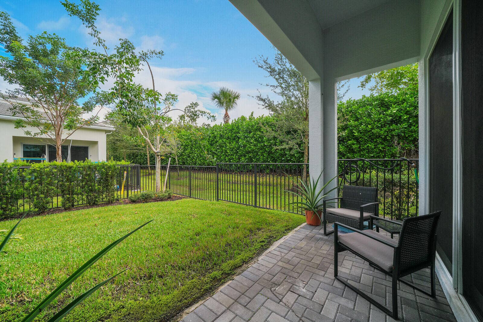 Enjoy serenity in your covered patio featuring paver flooring, enclosed backyard with manicured lawn, black metal fencing, and perimeter privacy hedges