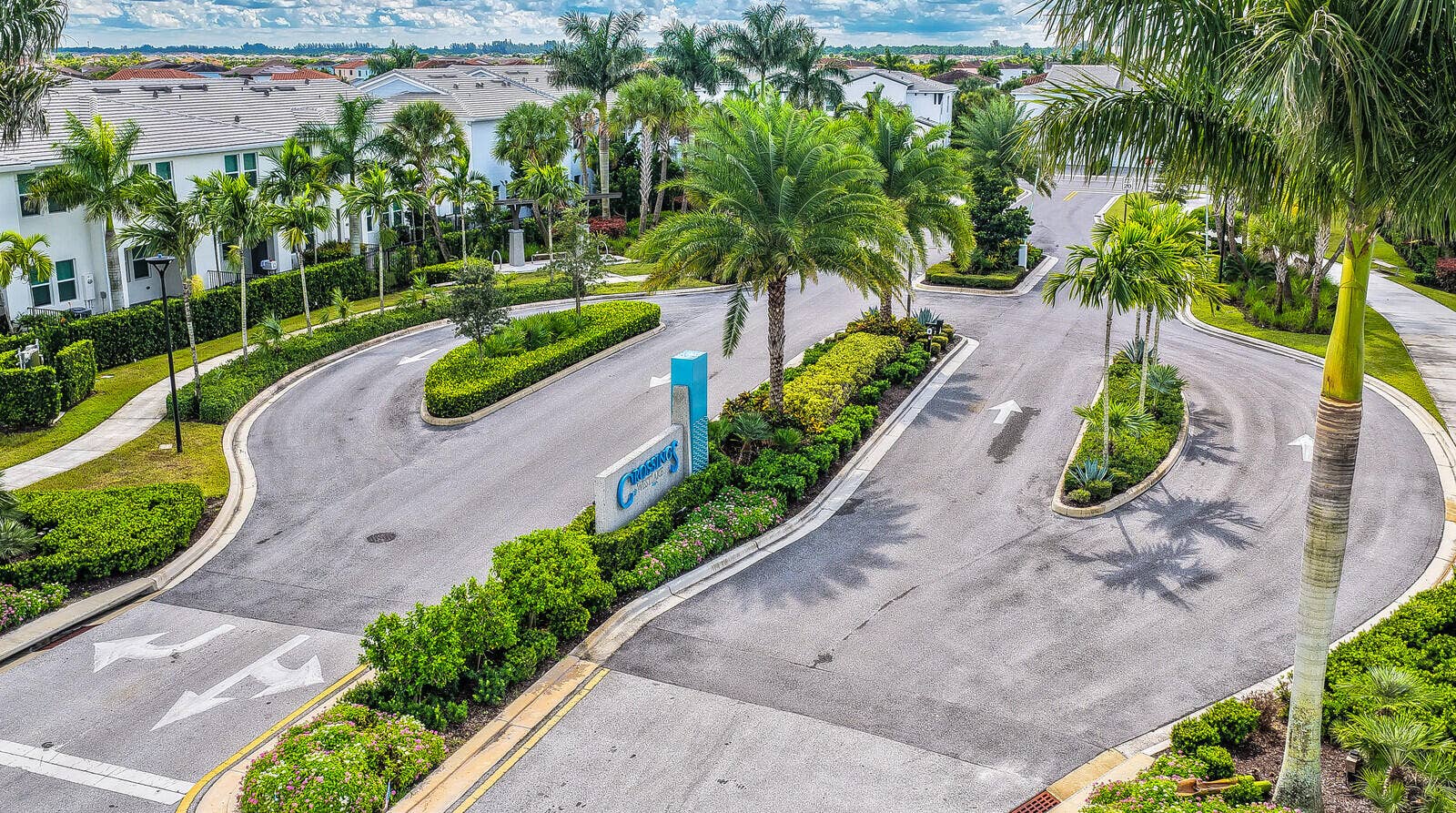 Gated entrance featuring landscaped medians, mature palm trees, paved roadways, and a prominent community monument sign