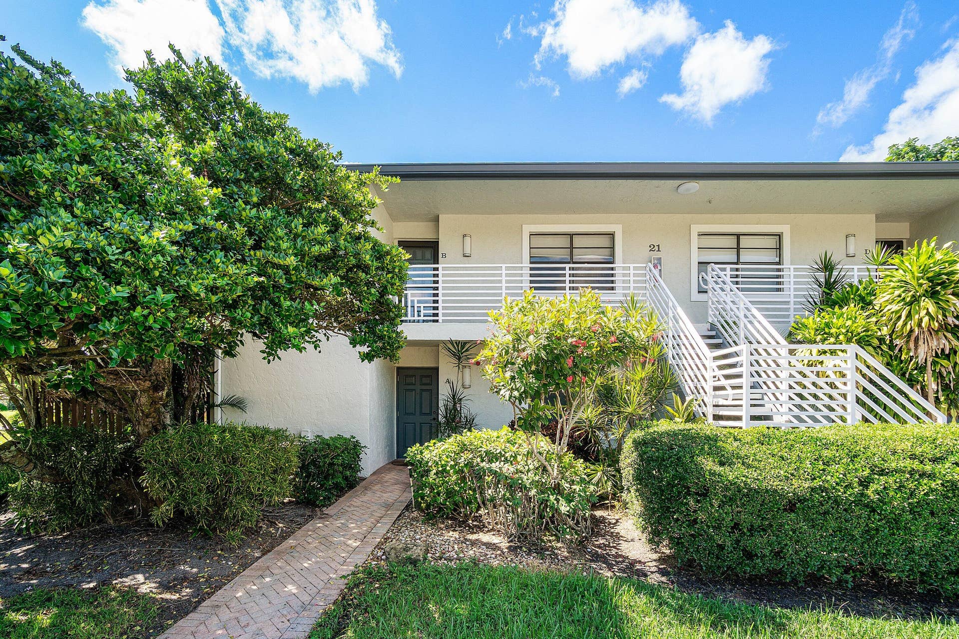 21 Southport Lane, #B, Boynton Beach, FL 33436 Multi-level building exterior featuring white stucco siding, a second-story balcony with horizontal slat railing, a concrete staircase, and a brick paver walkway