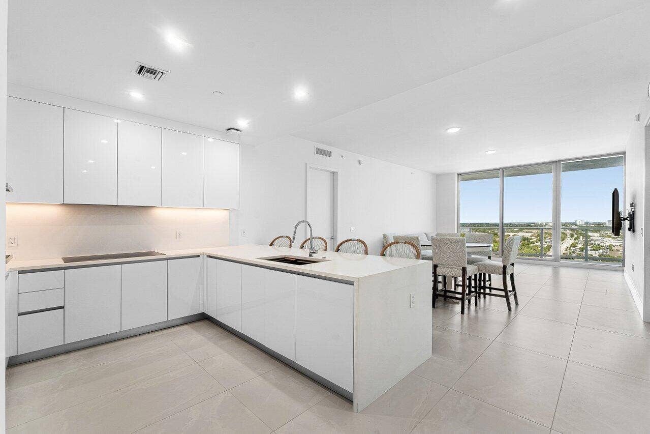 Minimalist kitchen featuring high-gloss white cabinetry, solid-surface countertops, a center island with an integrated sink, under-cabinet lighting, and large-format tile flooring