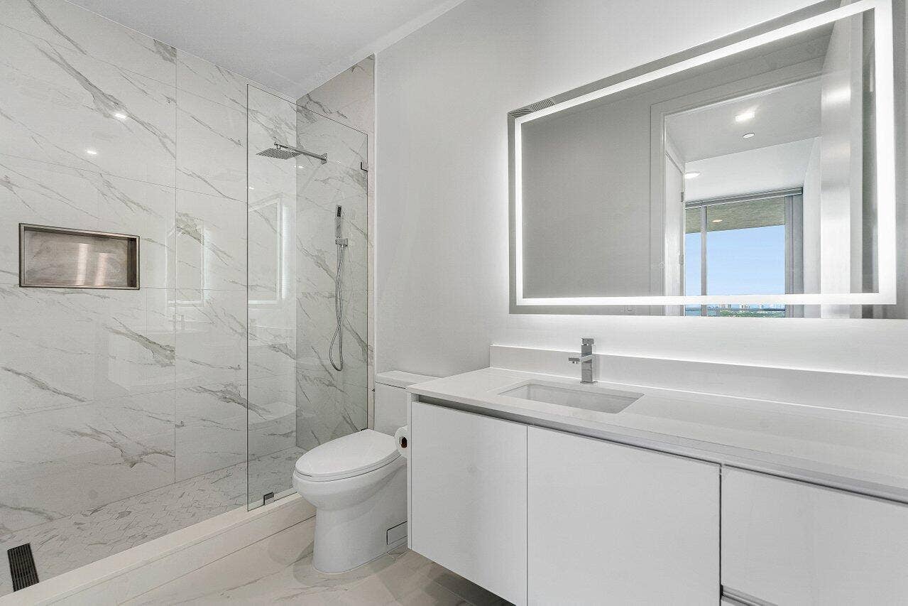 Bathroom featuring floor-to-ceiling stone-look tiled shower with integrated niche and glass enclosure, illuminated vanity mirror, and flat-panel cabinetry