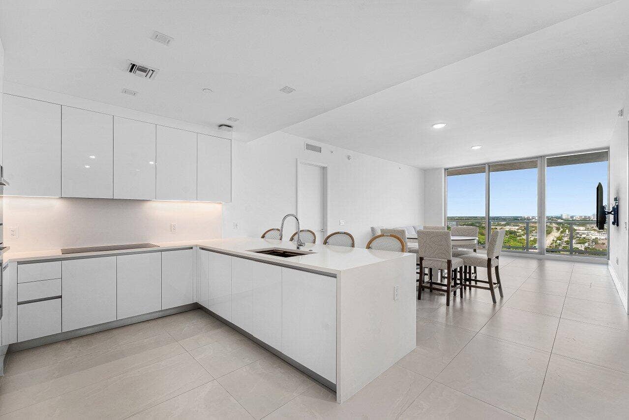 Modern kitchen featuring flat-panel white cabinetry, quartz countertops, a central island with an integrated sink, recessed lighting, and expansive floor-to-ceiling windows