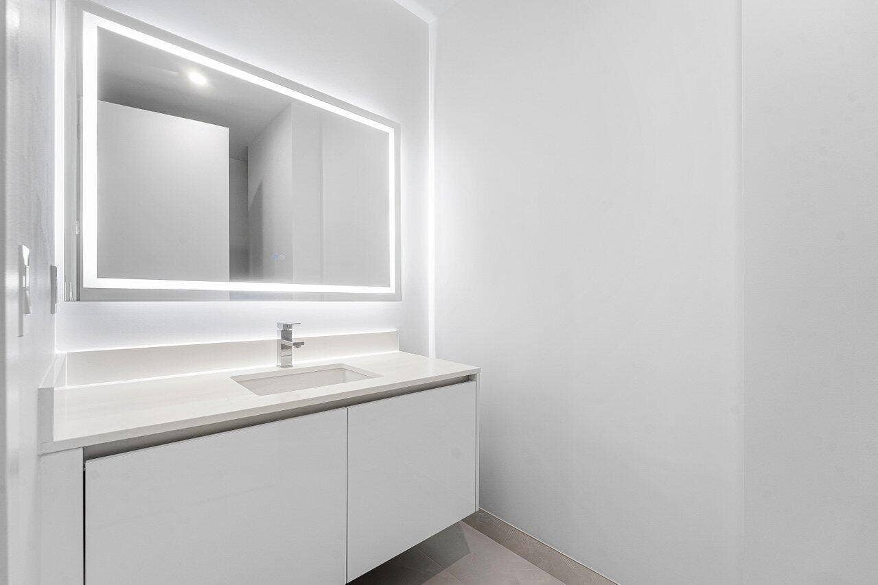 Floating vanity featuring flat-panel cabinetry, a light-toned countertop with an integrated sink, polished chrome faucet, and a large rectangular LED-backlit mirror