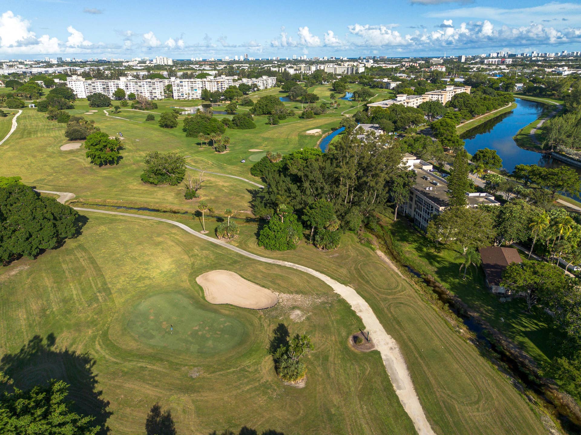 Aerial perspective of a golf course featuring manicured greens, sand bunkers, winding paths, mature trees, and a bordering waterway