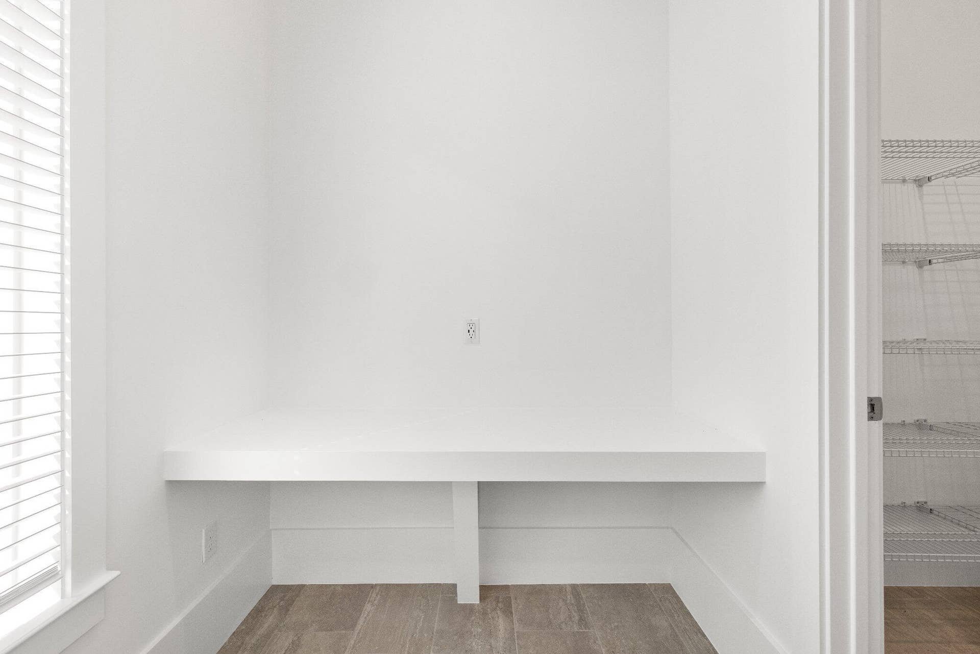 Built-in desk with a crisp white finish, horizontal blinds, wood-finish flooring, and white wire shelving