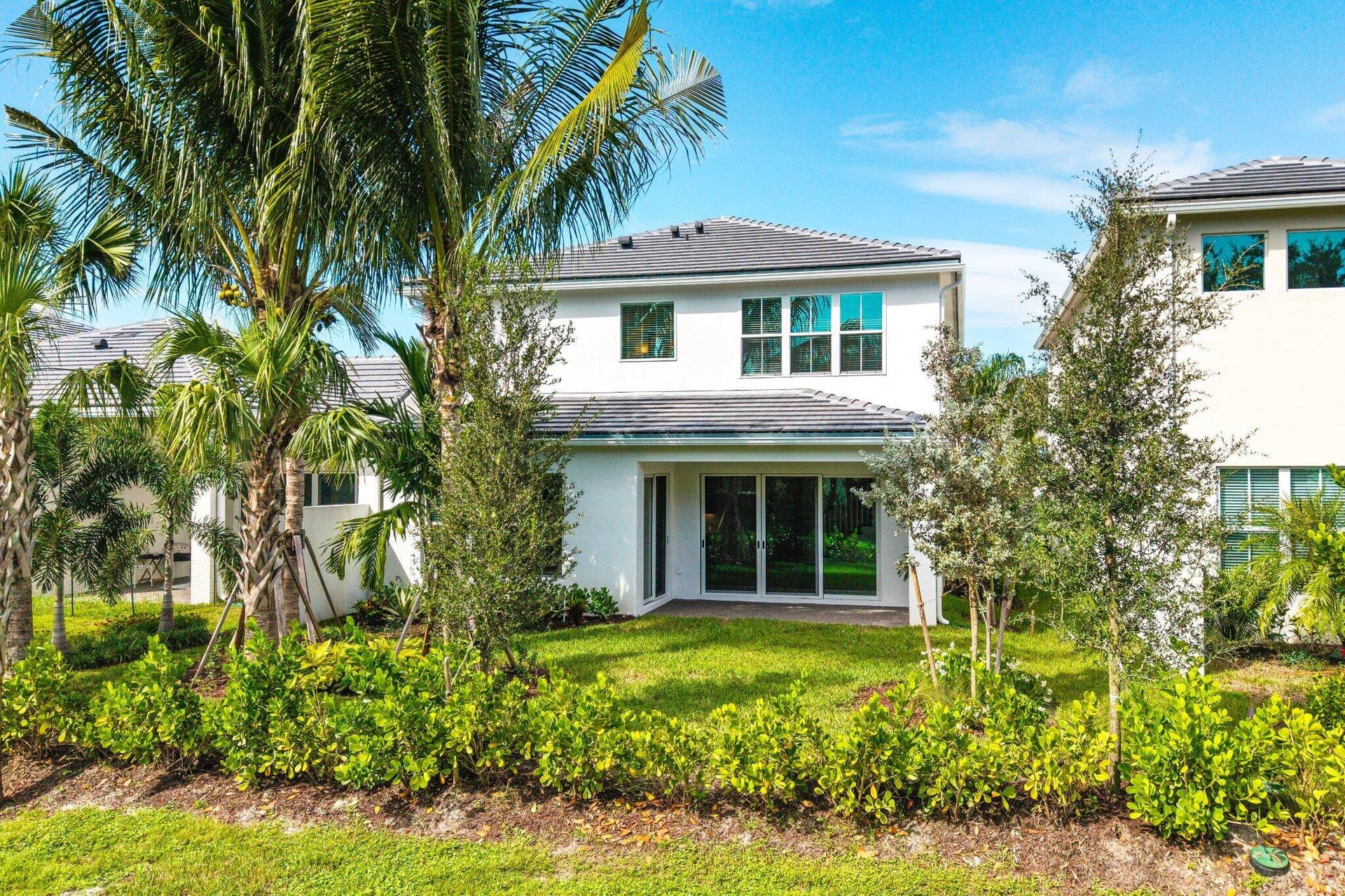 Two-story exterior featuring white stucco siding, a grey tile roof, multi-pane windows, and sliding glass doors with a grass lawn and established perimeter landscaping