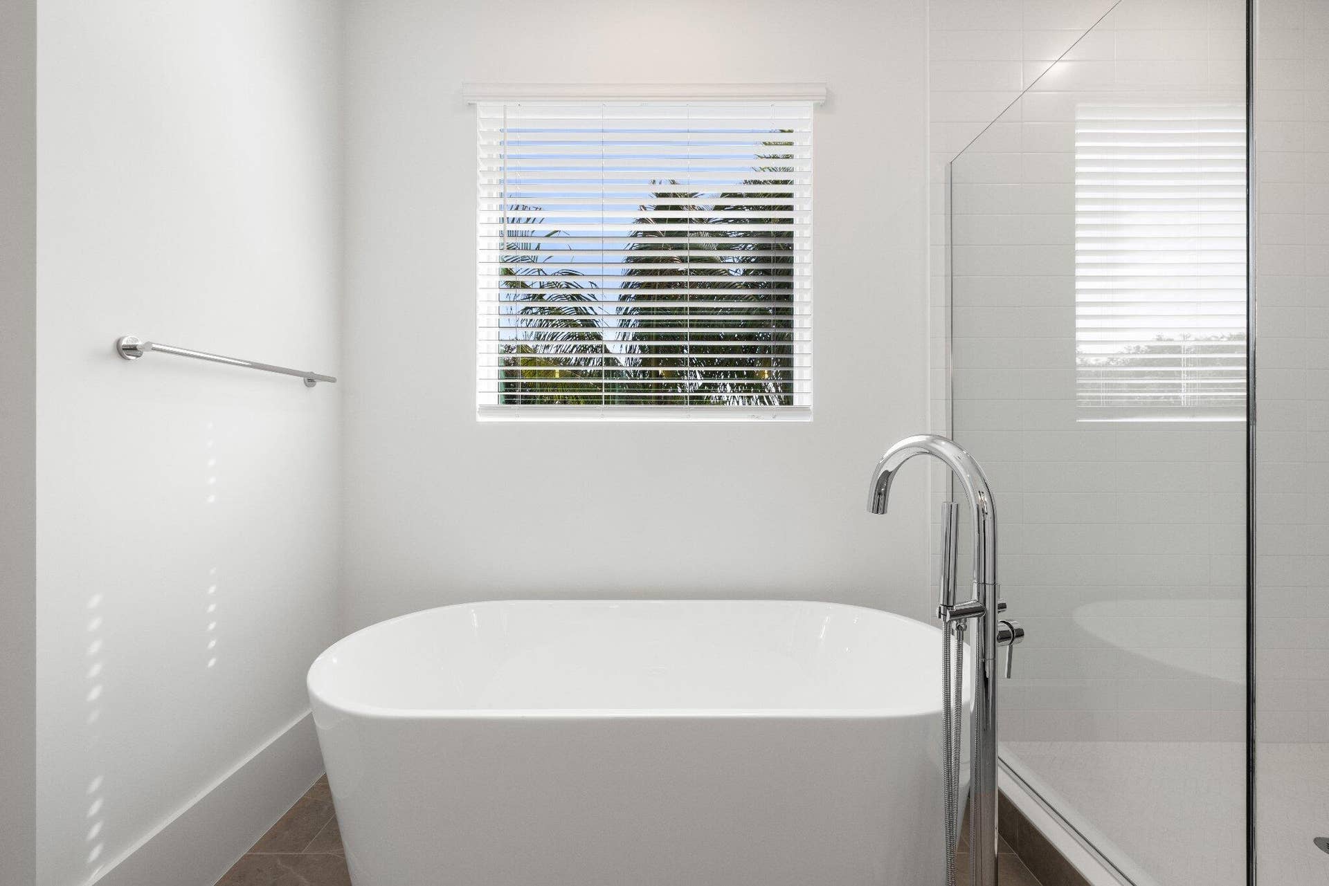 Freestanding soaking tub with polished chrome floor-mounted faucet, glass-enclosed shower, tile flooring, and window with slatted blinds