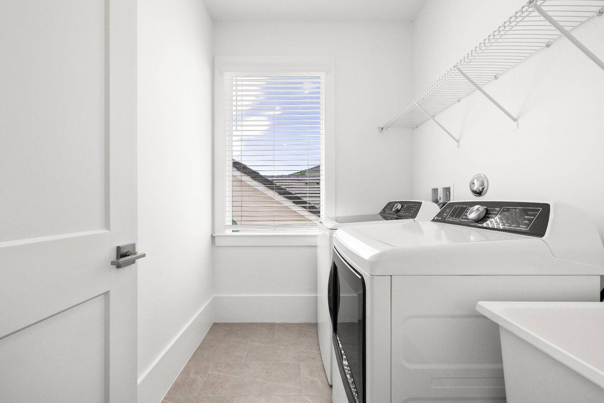 Utility room featuring a window with horizontal blinds, neutral tile flooring, and overhead wire shelving