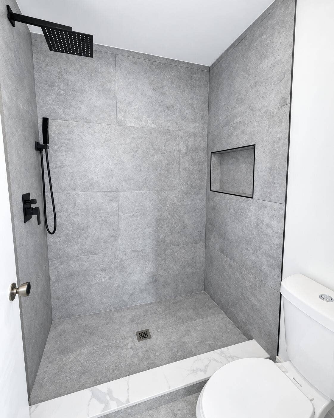 Walk-in shower featuring large-format grey tile, a matte black rainfall showerhead, a matching handheld wand, and a built-in recessed wall niche