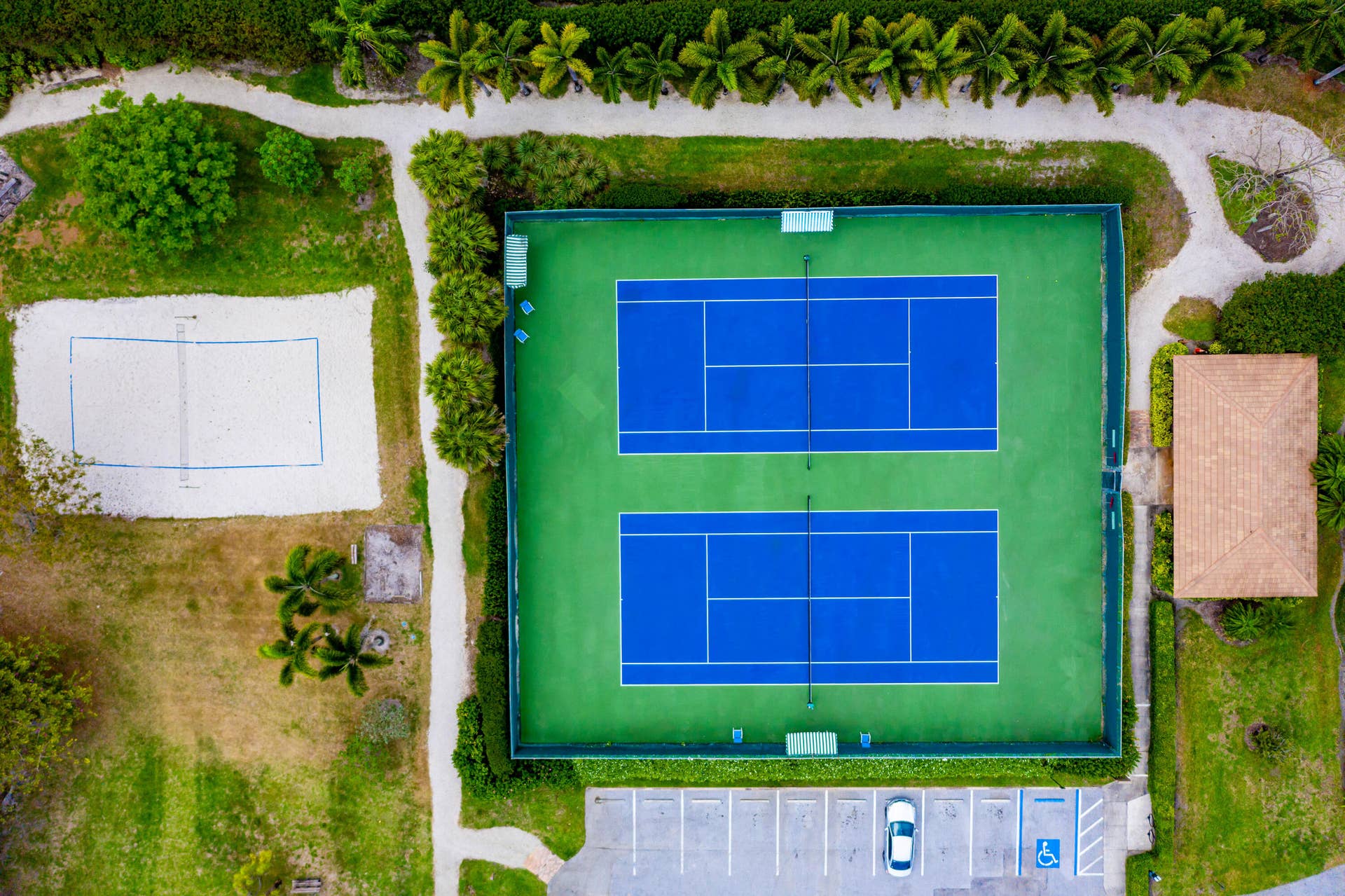 137 Clubhouse Boulevard, Atlantis, FL 33462 Dual tennis courts with fencing, adjacent sand court, and paved parking area surrounded by mature landscaping