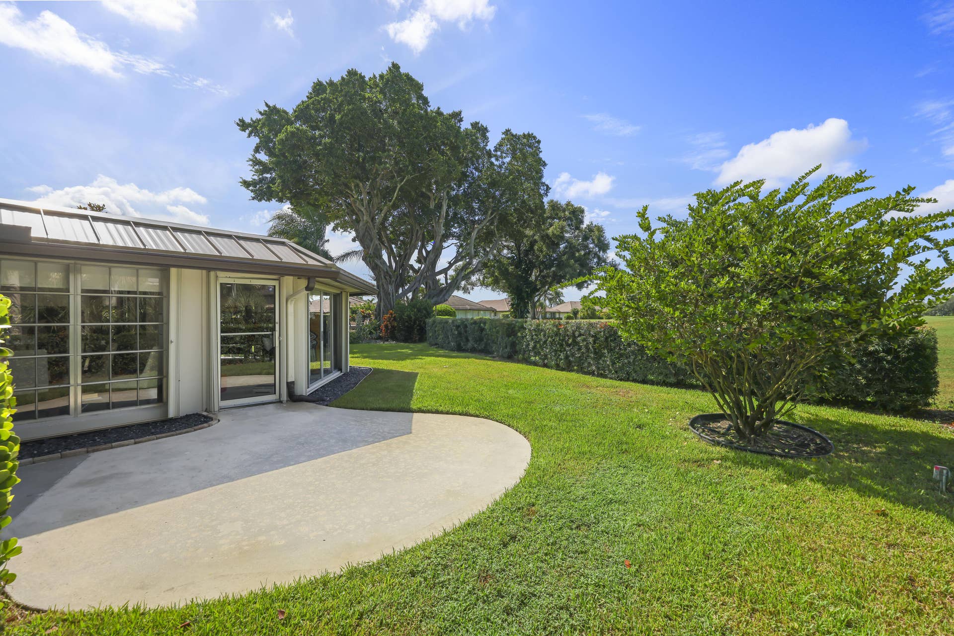 137 Clubhouse Boulevard, Atlantis, FL 33462 Rear exterior featuring a concrete patio, metal roofing, expansive manicured lawn, established landscaping, and mature trees