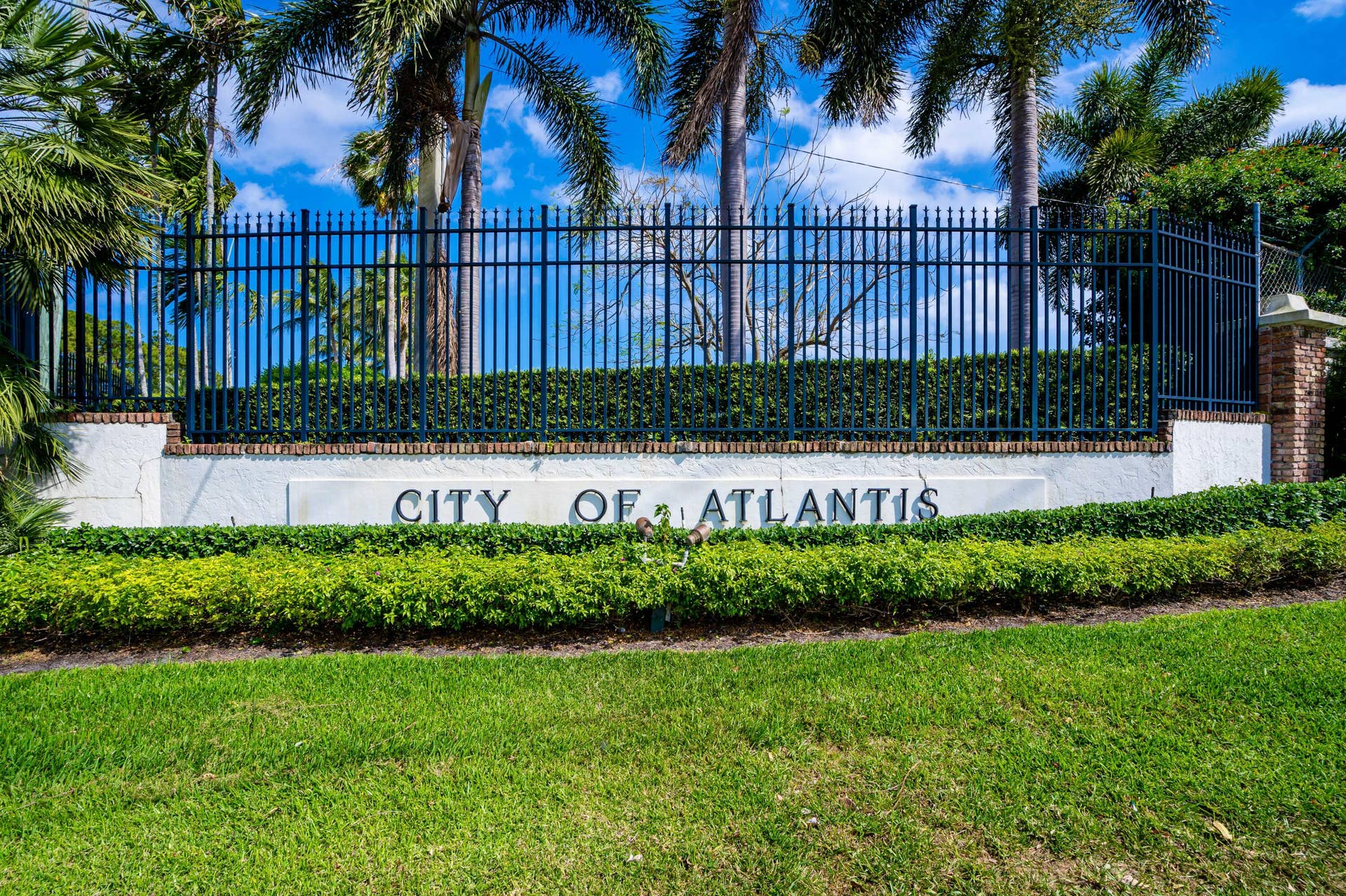 137 Clubhouse Boulevard, Atlantis, FL 33462 Community entrance featuring a concrete masonry wall, black metal fencing, brick pillar accents, and professionally manicured landscaping