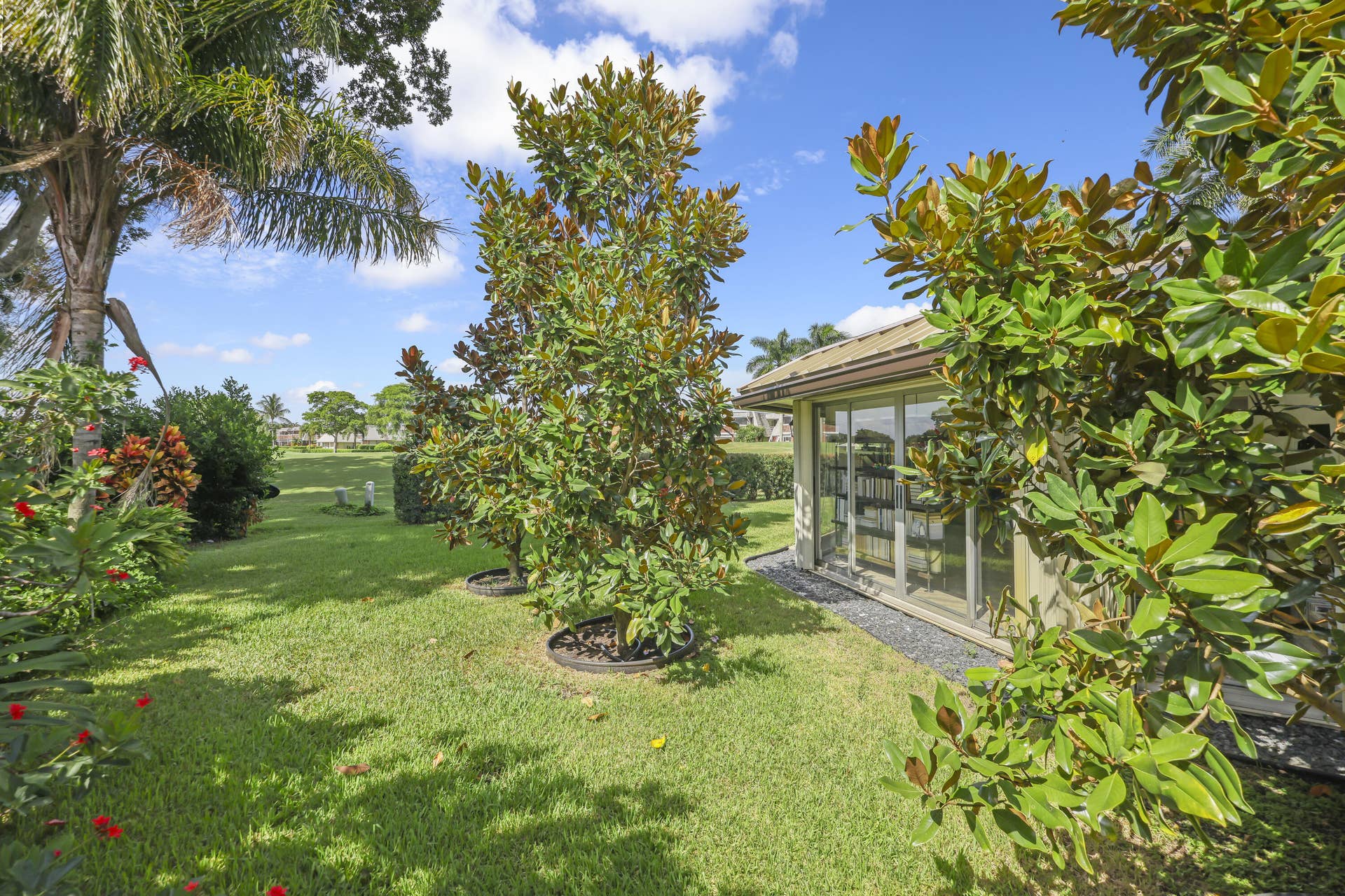 137 Clubhouse Boulevard, Atlantis, FL 33462 Expansive lawn with mature landscaping, including established trees and shrubs, featuring a structural exterior with floor-to-ceiling sliding glass doors and gravel perimeter accent