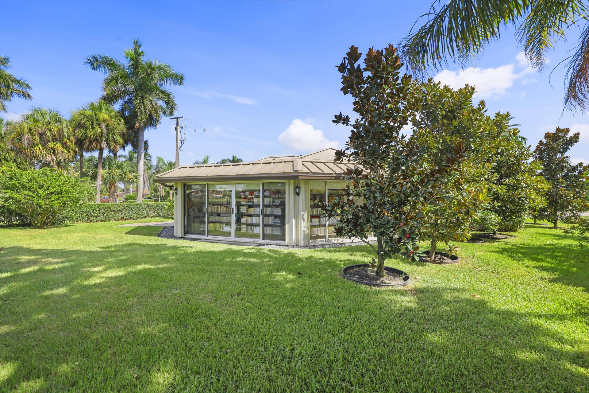 137 Clubhouse Boulevard, Atlantis, FL 33462 Standalone structure featuring a metal hip roof, sliding glass wall panels, exterior wall-mounted lighting, and expansive landscaped grounds with mature trees