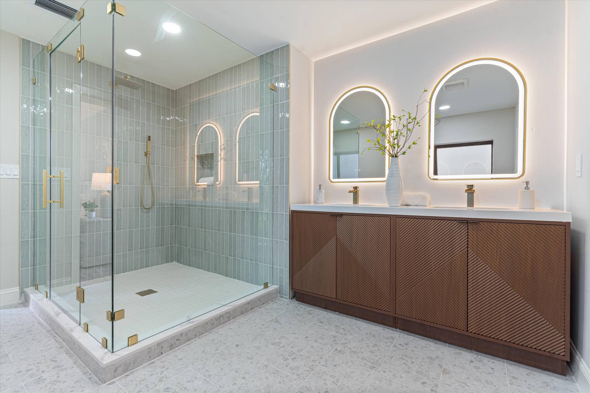 6111 WHITE OAK Lane, Tamarac, FL 33319 Double vanity featuring textured wood cabinetry, stone countertops, integrated arched LED-backlit mirrors, gold-finish hardware, and a glass-enclosed shower with vertical wall tile