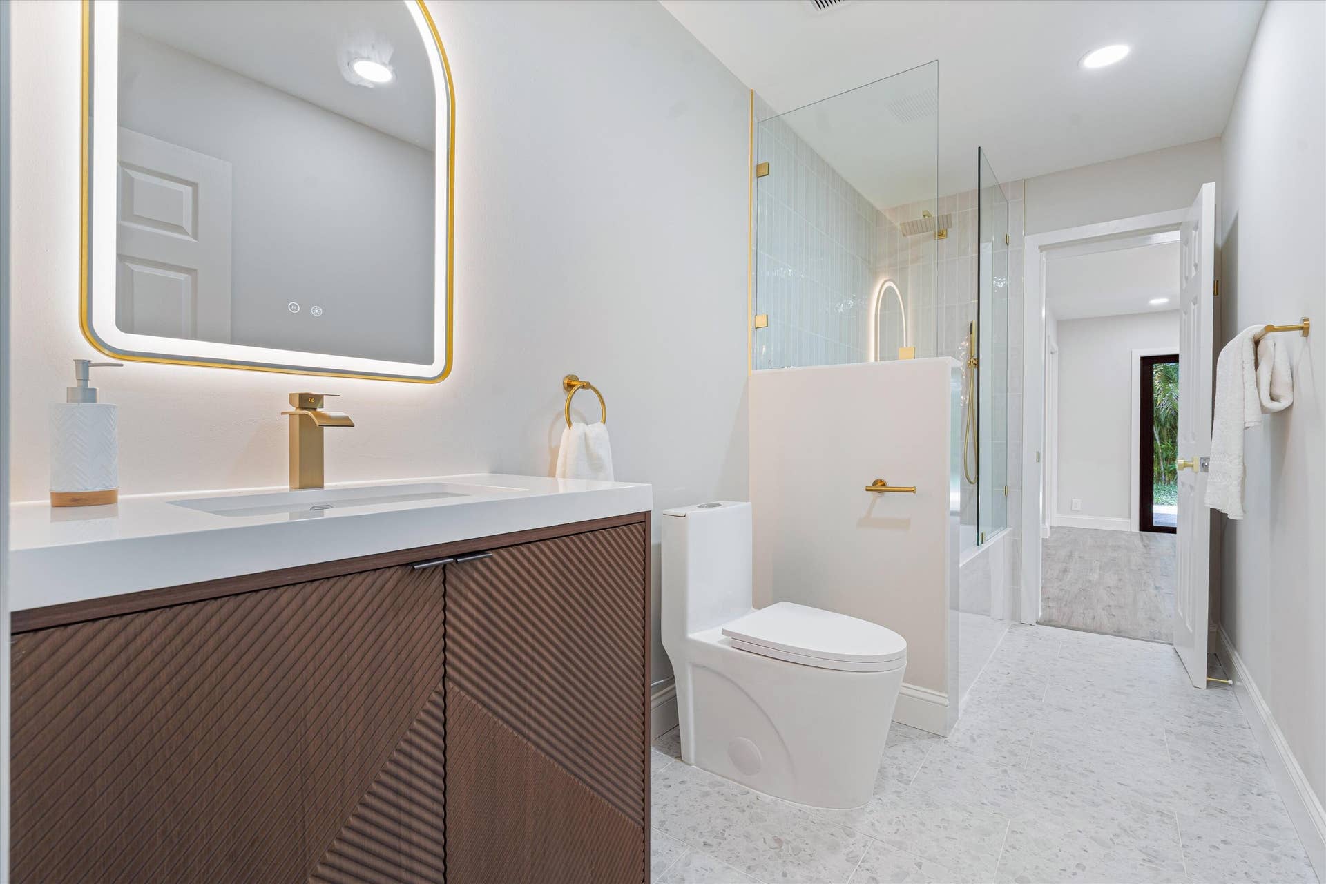 6111 WHITE OAK Lane, Tamarac, FL 33319 Floating vanity with a textured wood finish, solid surface countertop, brass-toned hardware, arched LED-backlit mirror, and glass-enclosed shower