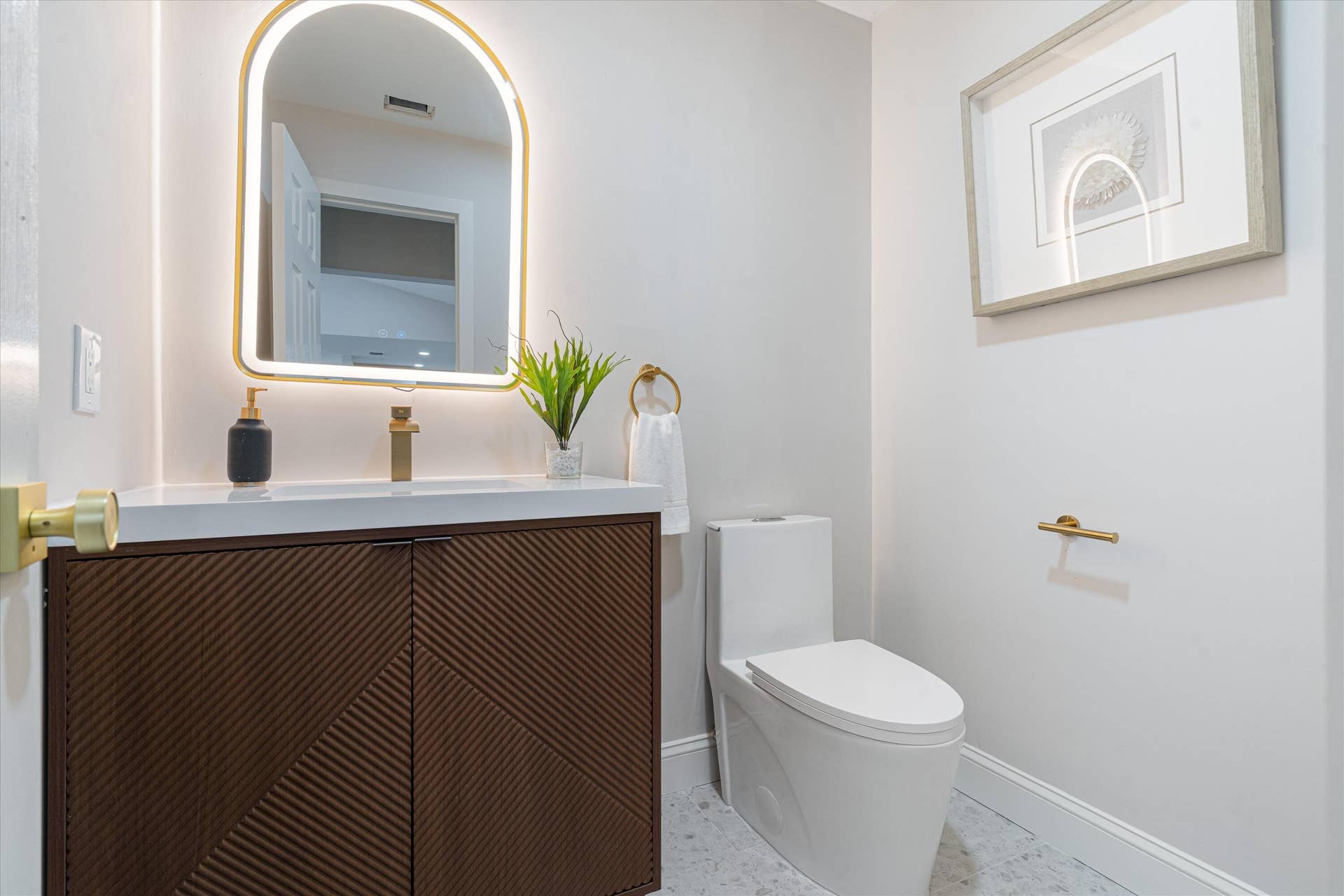6111 WHITE OAK Lane, Tamarac, FL 33319 Modern vanity with textured wood-finish cabinet doors, white surface, arched backlit mirror, brass-finish fixtures, and a one-piece dual-flush toilet
