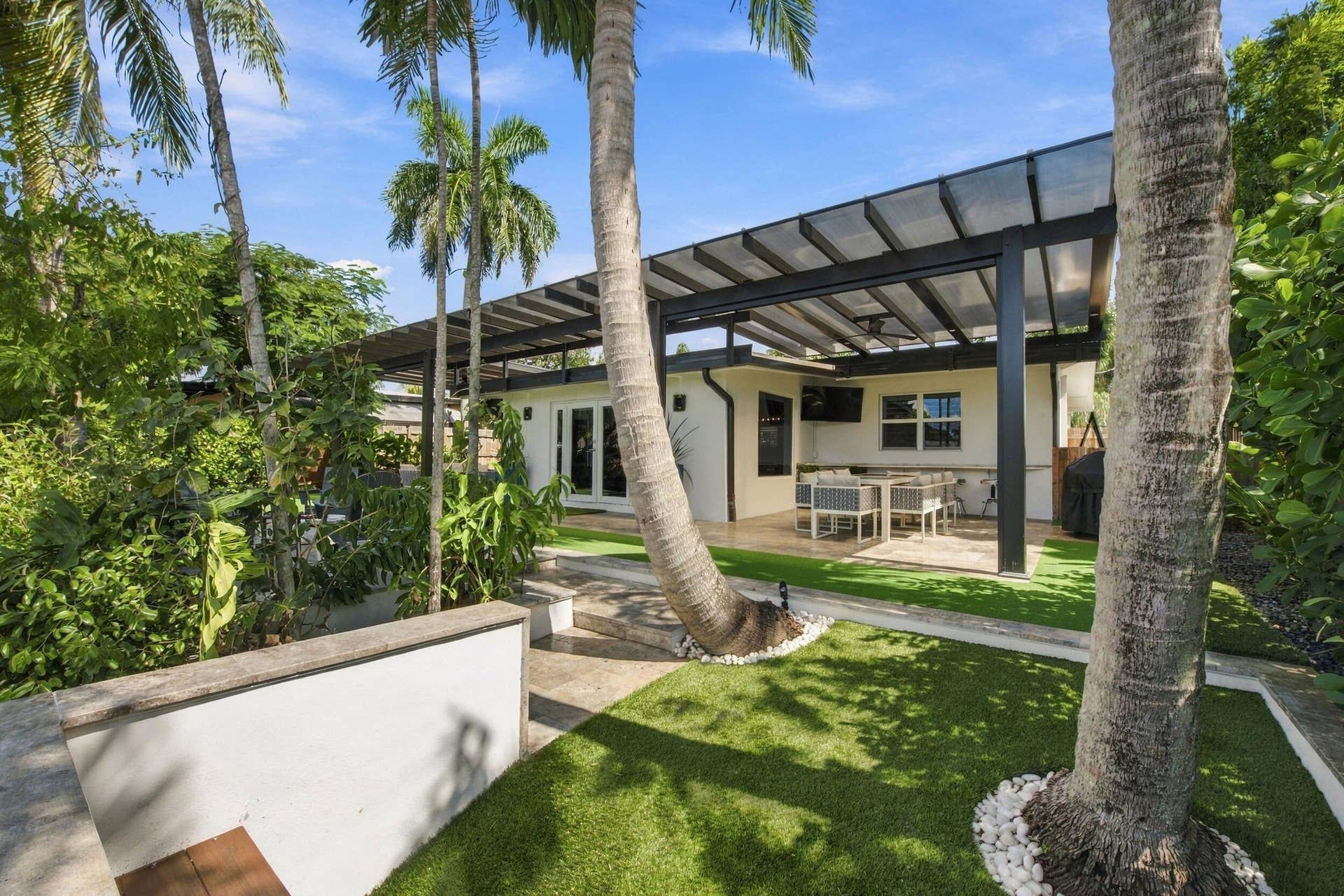 2449 KEY LARGO Lane, Fort Lauderdale, FL 33312 Outdoor living space featuring a modern pergola with a semi-translucent roof, stone patio flooring, synthetic lawn, and mature palm trees