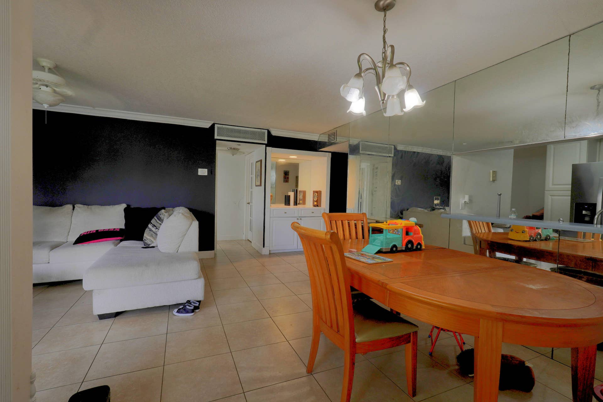 9853 NW 3RD Court, #7, Plantation, FL 33324 Open living and dining area featuring large-format tile flooring, a multi-arm polished metal chandelier, mirrored wall paneling, and a built-in white cabinetry unit with recessed lighting