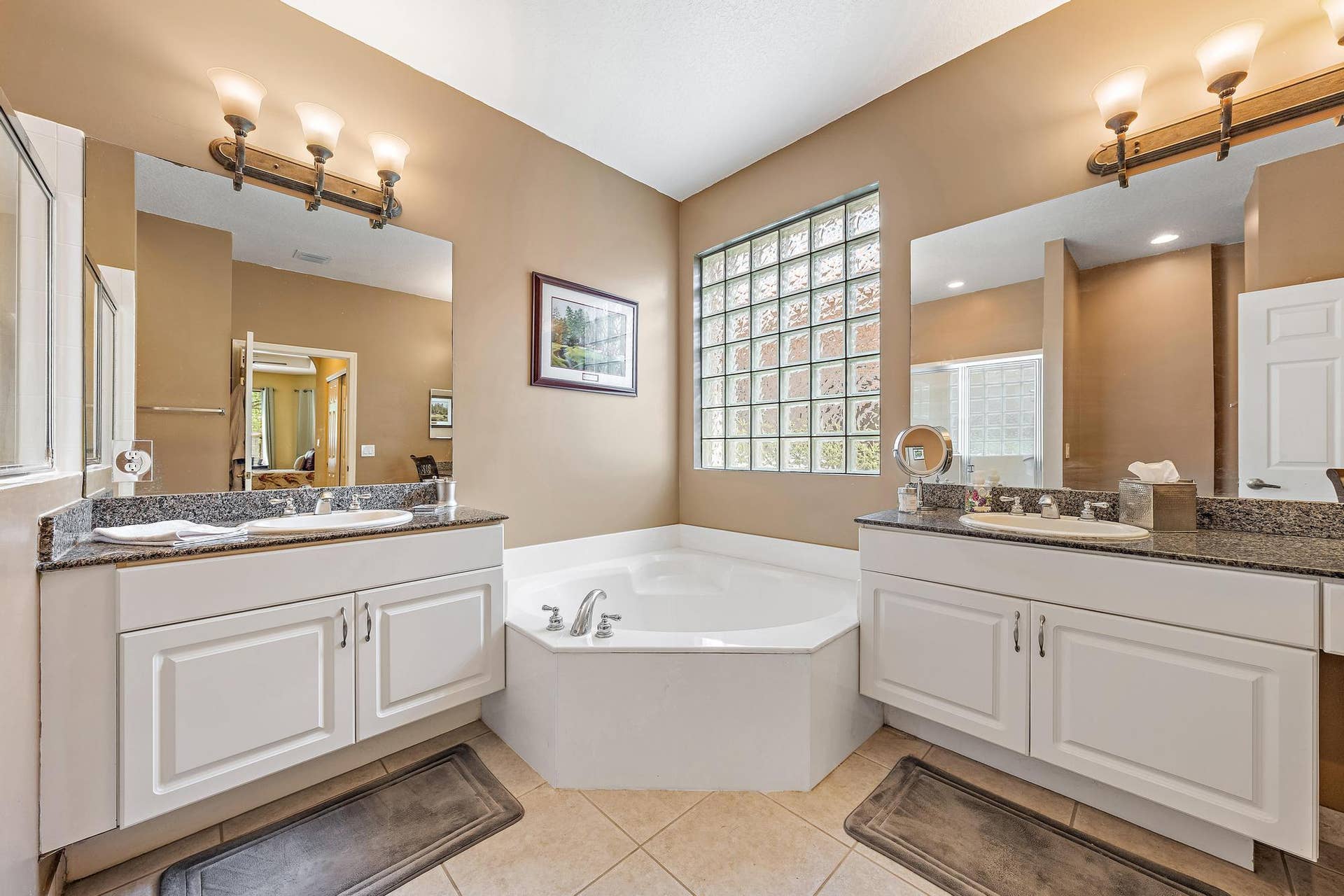 Dual white vanity cabinets featuring stone-surface countertops, integrated oval sinks, glass block accent window, and a built-in corner soaking tub