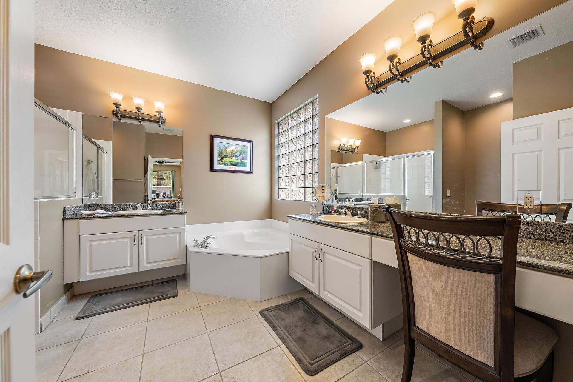 Bathroom featuring dual white cabinetry with stone-surface countertops, soaking tub, glass-block window, and ceramic tile flooring