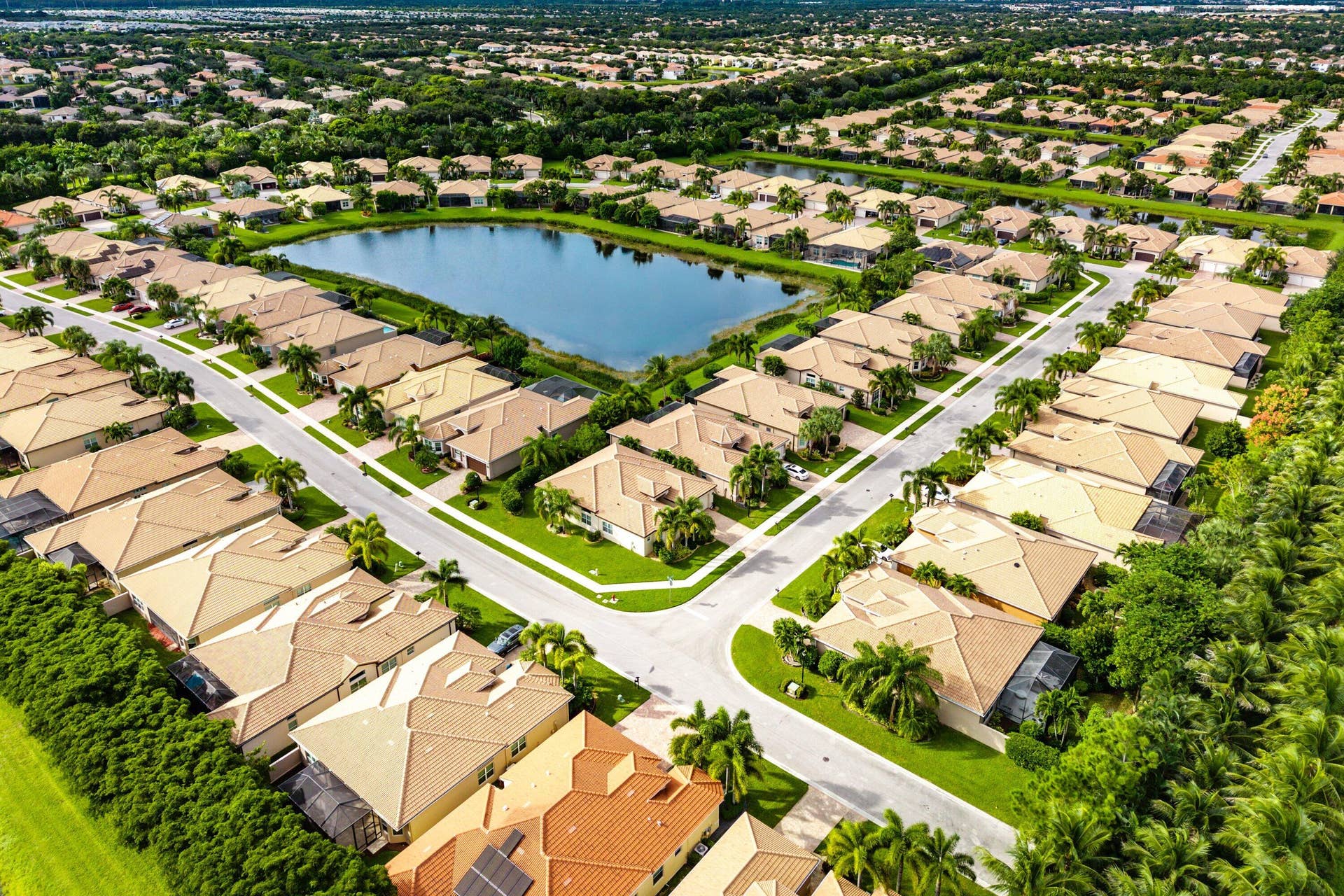 Aerial perspective of a planned community featuring a central lake, paved street networks, tile-roof residential structures, and mature palm trees