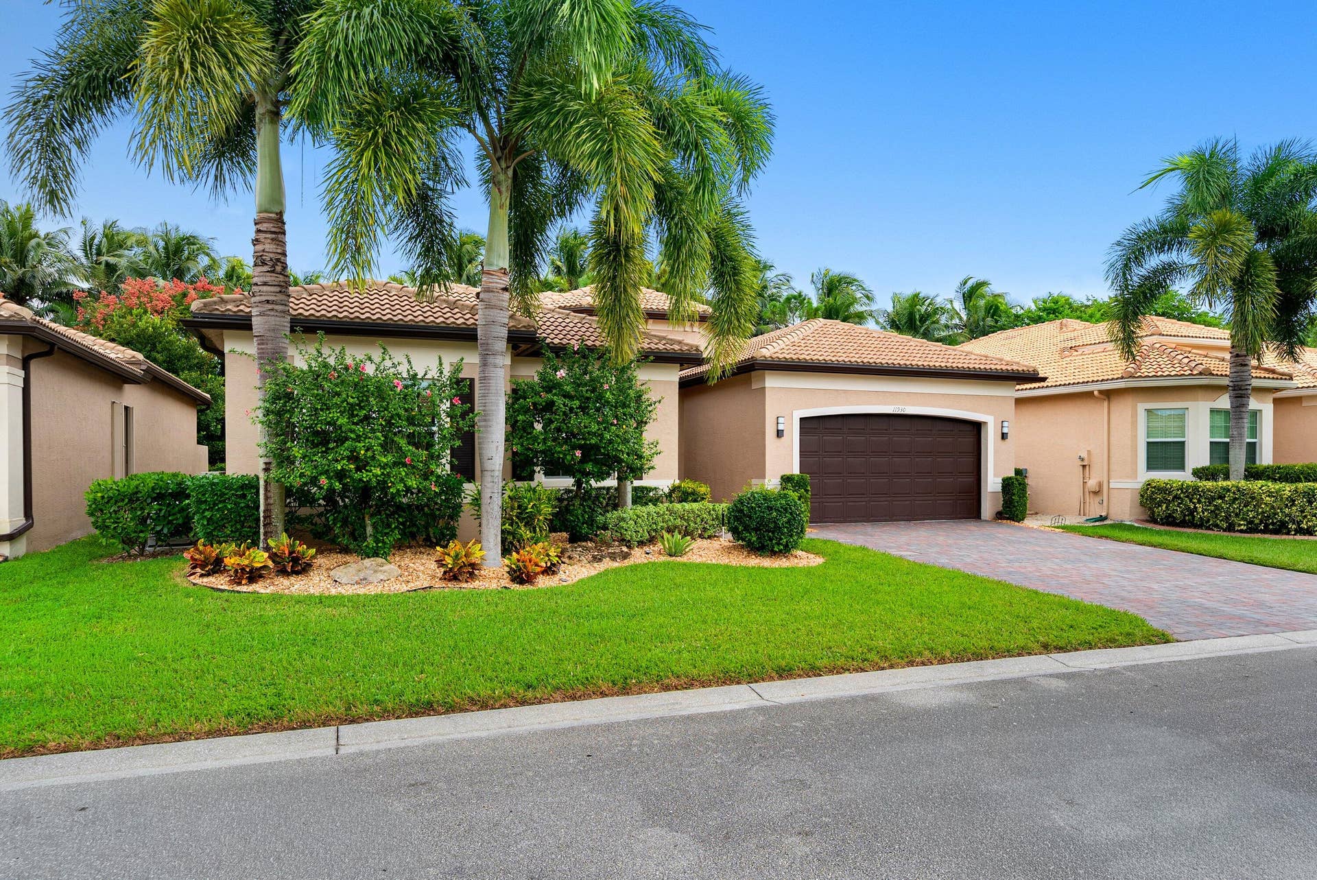 Stucco exterior residence featuring a tile roof, paved driveway, two-car garage, and established landscaping with mature palm trees