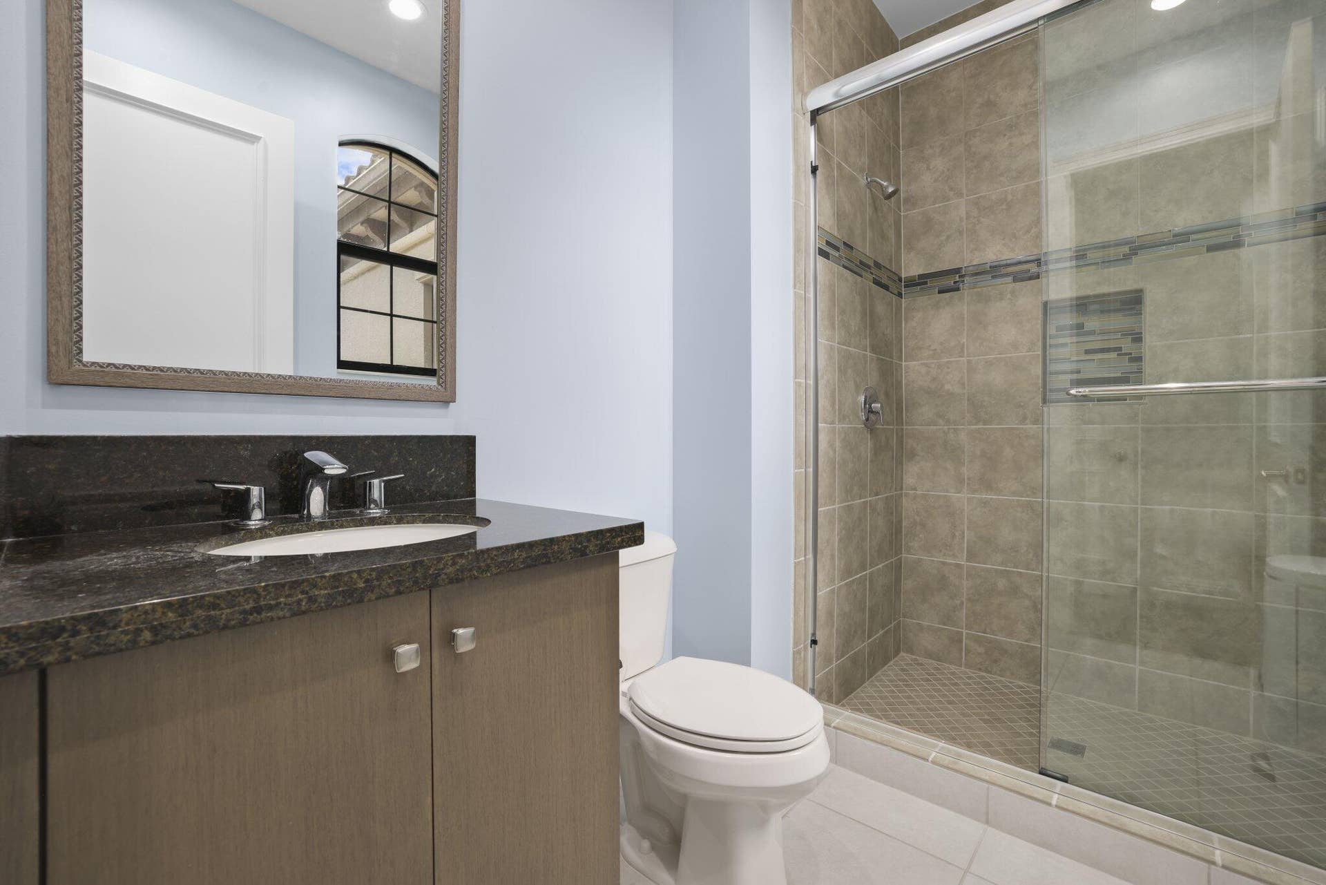 Granite-topped vanity with undermount sink, chrome faucet, framed mirror, and glass-enclosed shower with tiled walls and mosaic accent