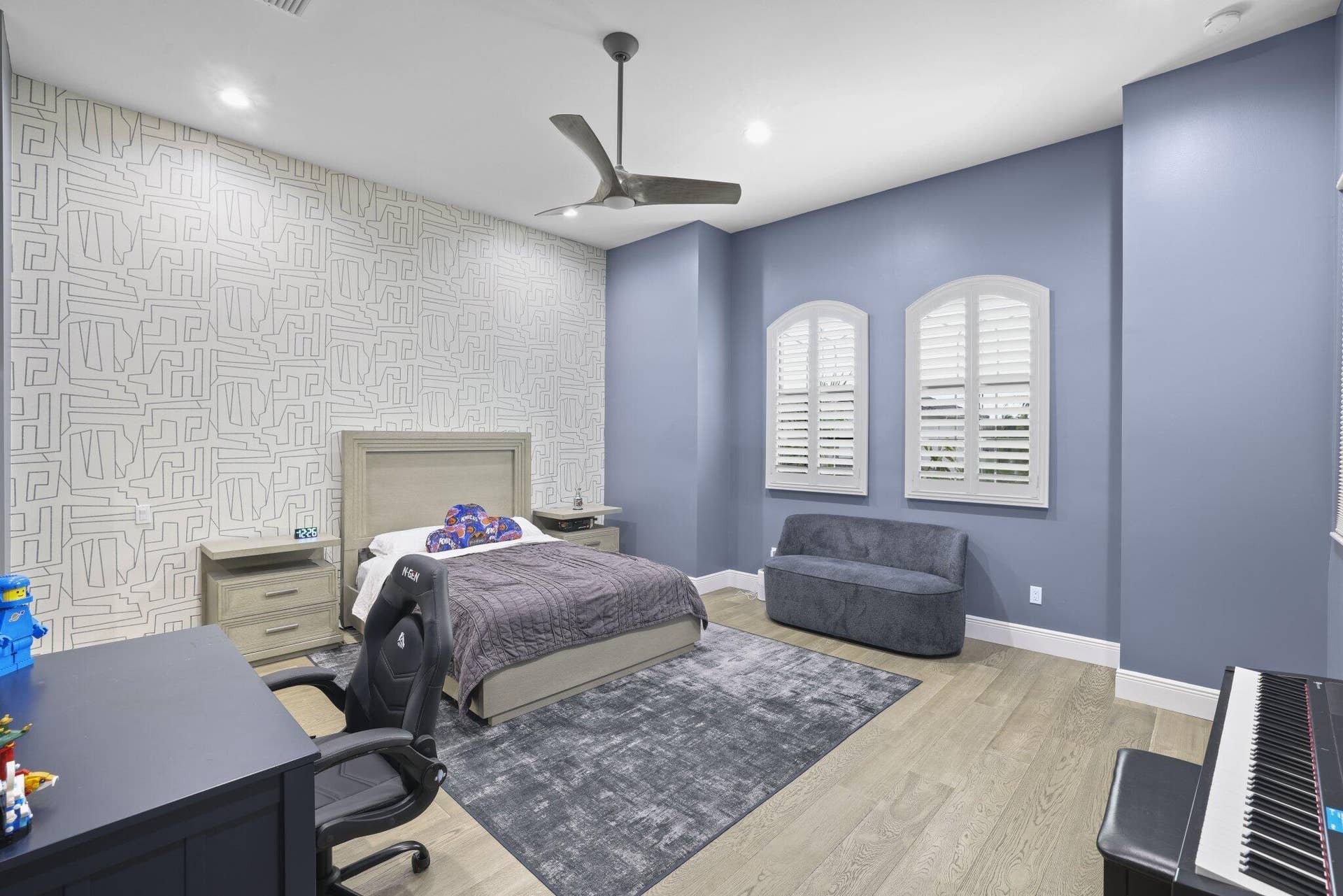 Light wood-finish flooring, accent wall with patterned wallpaper, arched windows with plantation shutters, ceiling fan, and recessed lighting