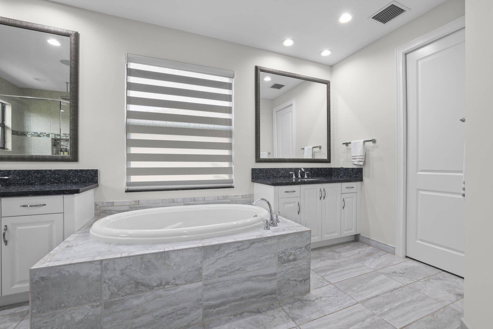 Built-in soaking tub with a tiled surround, dual vanities with dark countertops, and tiled flooring