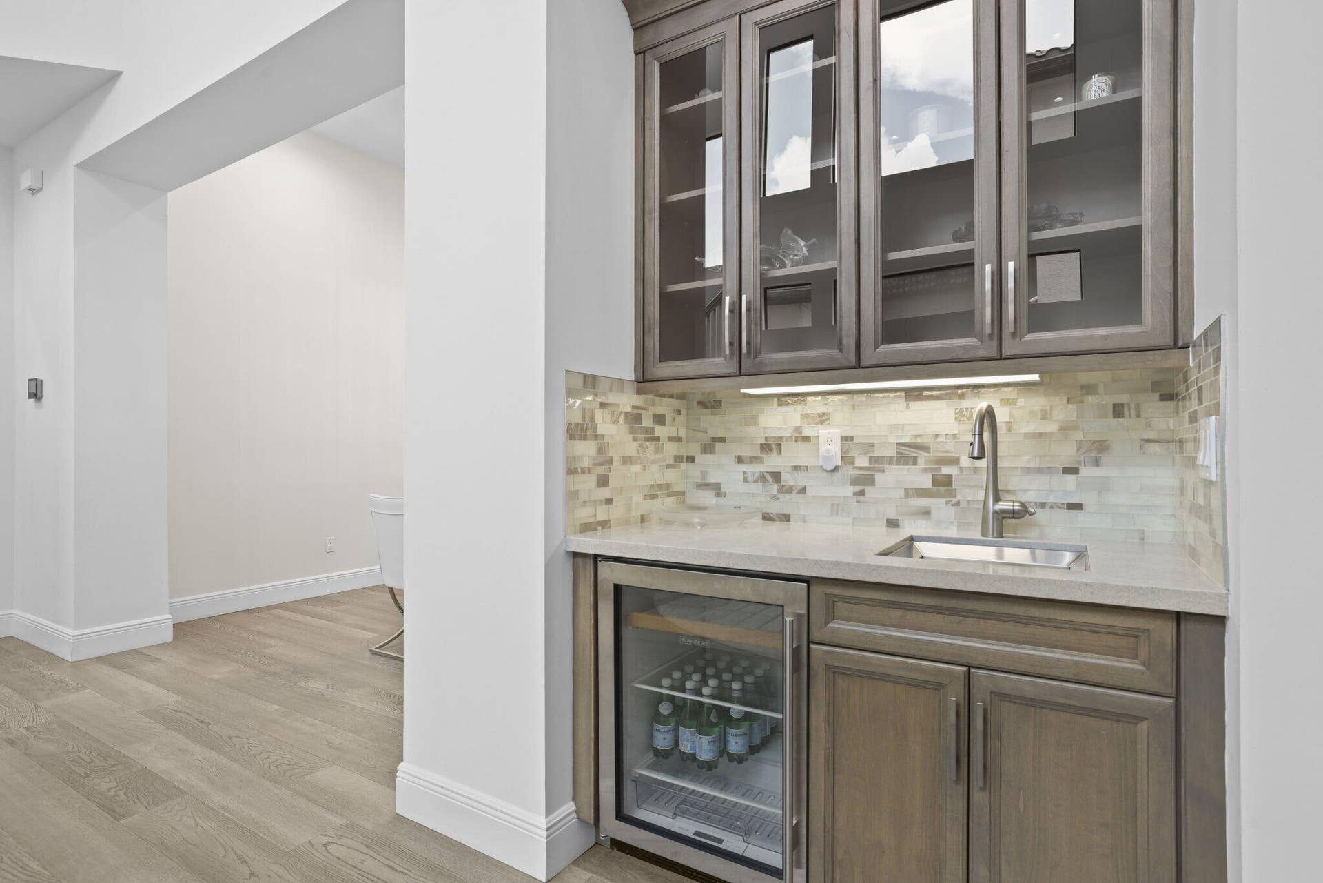Built-in wet bar featuring gray cabinetry, glass-front upper cabinets, light-toned countertop, mosaic tile backsplash, and a beverage refrigerator