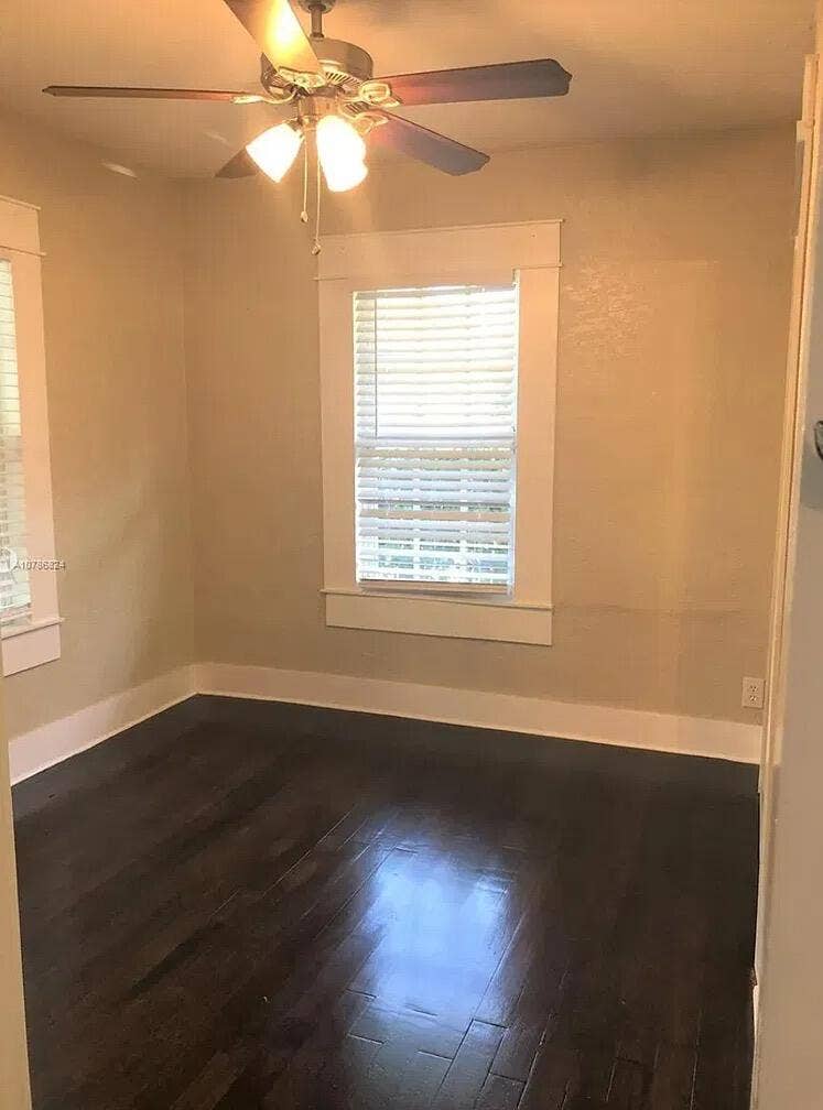 Interior room featuring dark wood-finish flooring, neutral wall paint, white millwork window trim, and a ceiling fan with integrated lighting