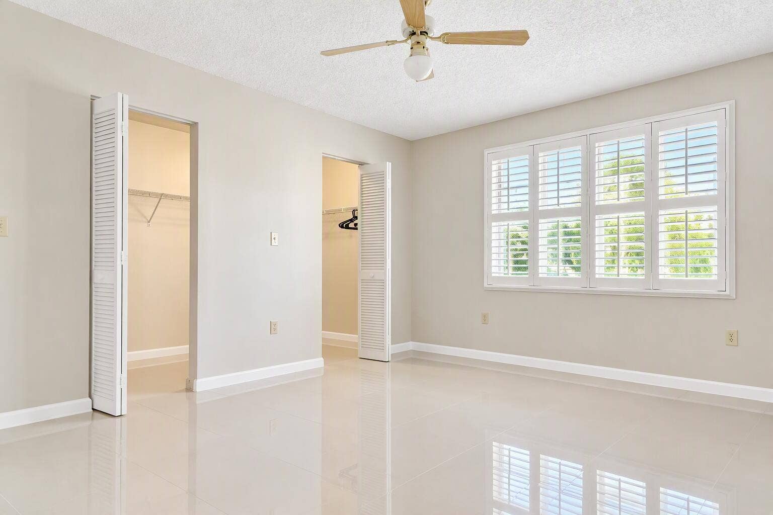 Neutral-toned interior featuring polished tile flooring, window shutters, dual closet entries with louvered doors, and a ceiling fan with light fixture