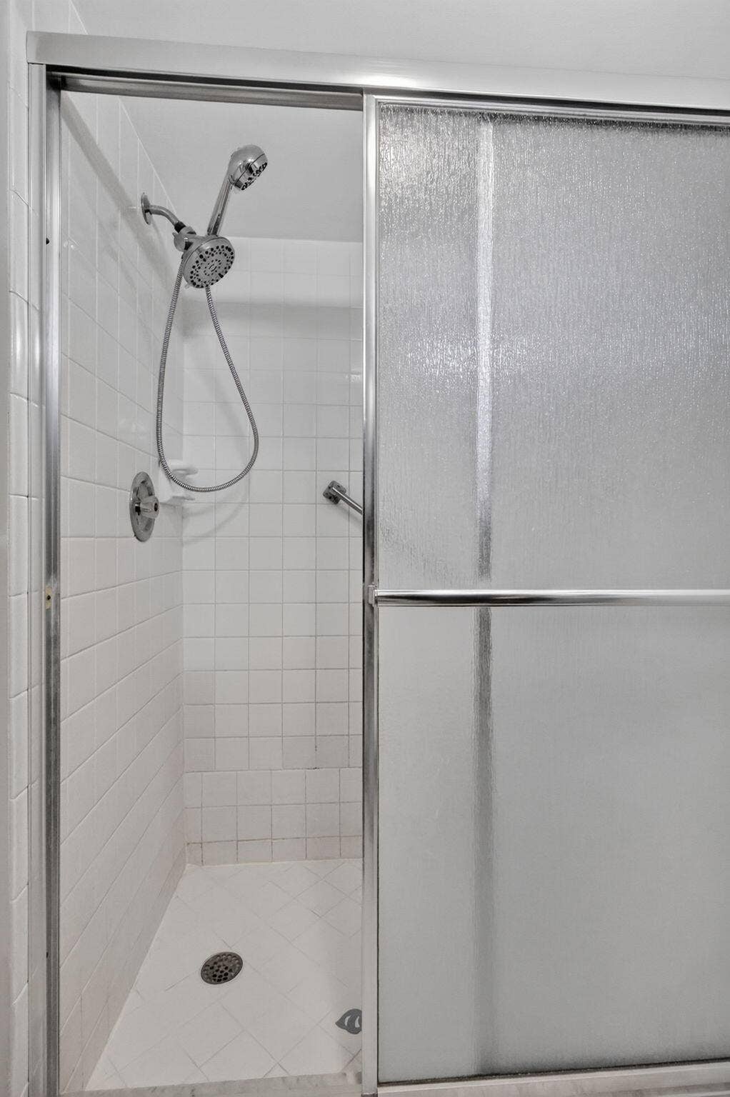14527 BONAIRE Boulevard, #109, Delray Beach, FL 33446 Shower enclosure featuring white tile walls, chrome-framed sliding textured glass doors, and a handheld shower fixture
