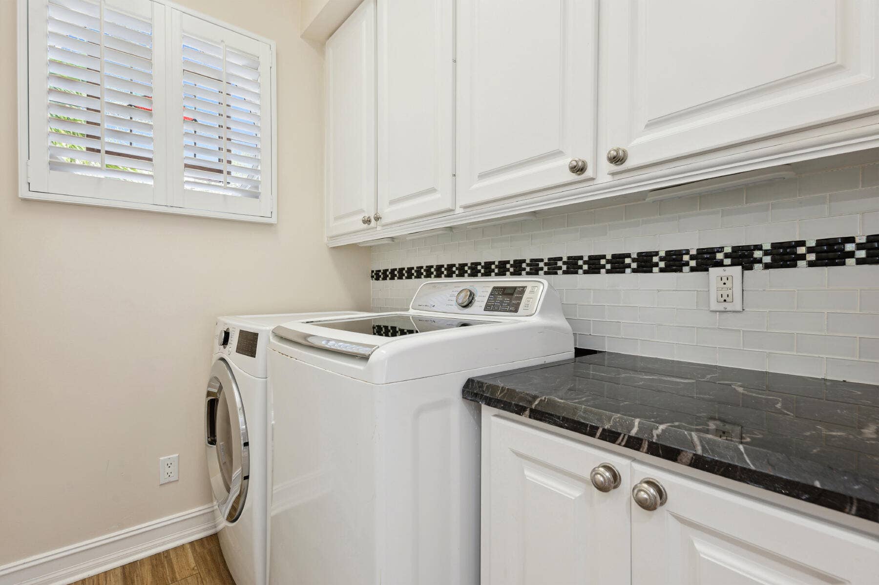 477 Capistrano Drive, Palm Beach Gardens, Palm Beach Gardens, FL 33410 Laundry area featuring white shaker-style cabinetry, dark stone-finish countertops, subway tile backsplash with decorative inset, and plantation shutters