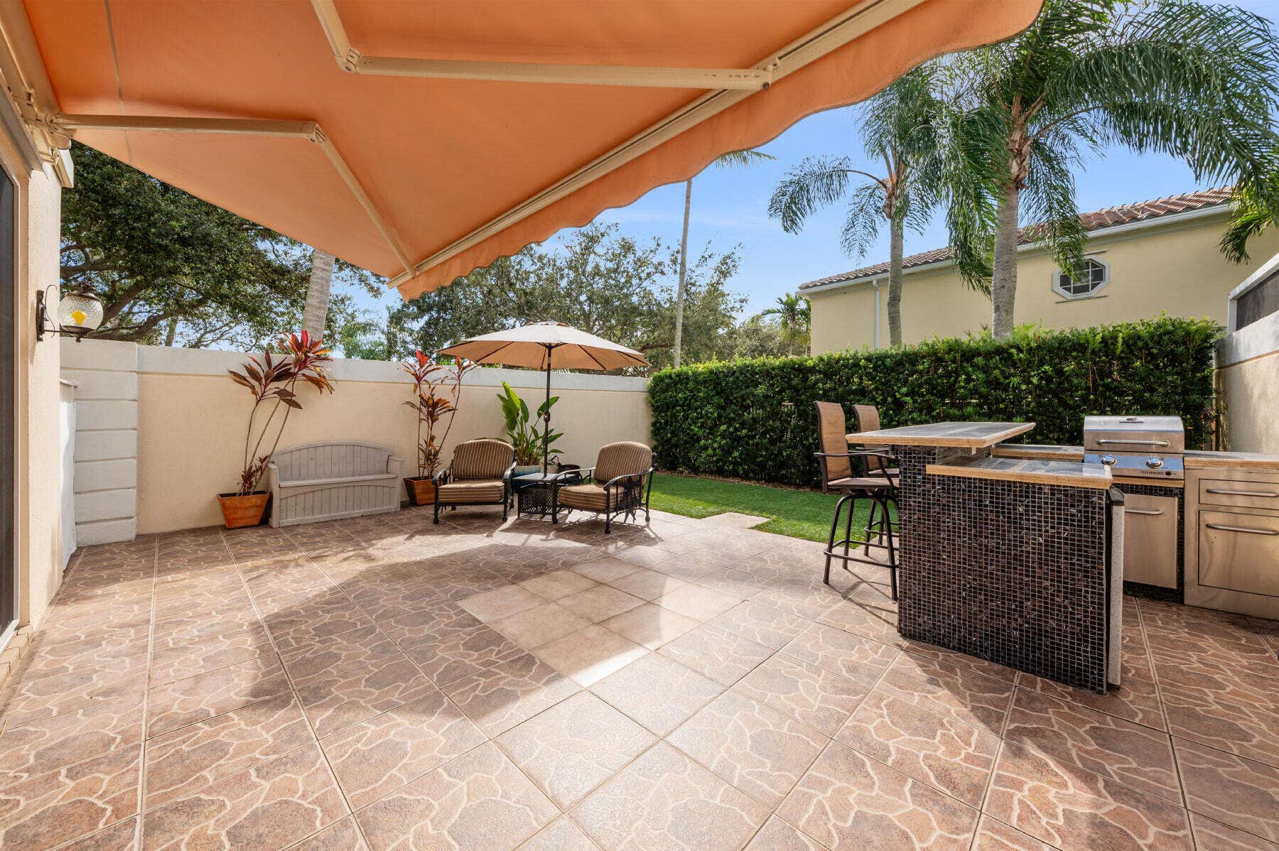 477 Capistrano Drive, Palm Beach Gardens, Palm Beach Gardens, FL 33410 Outdoor patio featuring a retractable awning, tiled flooring, and a built-in outdoor kitchen with stainless steel appliances and mosaic tile detailing, bordered by privacy walls and mature landscaping