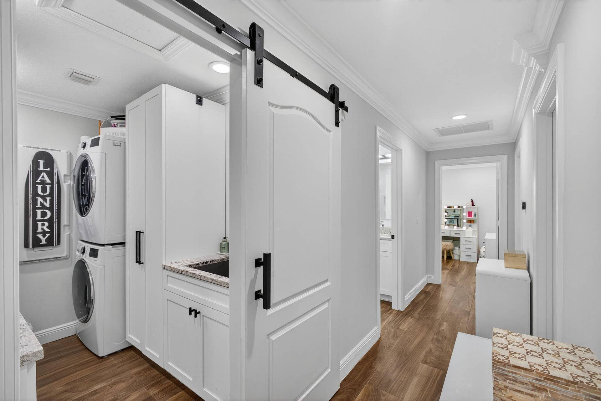 4628 Square Lake Drive, Palm Beach Gardens, FL 33418 Laundry room featuring a white sliding barn door with matte black hardware, stacked washer and dryer, custom white cabinetry, and wood-finish flooring