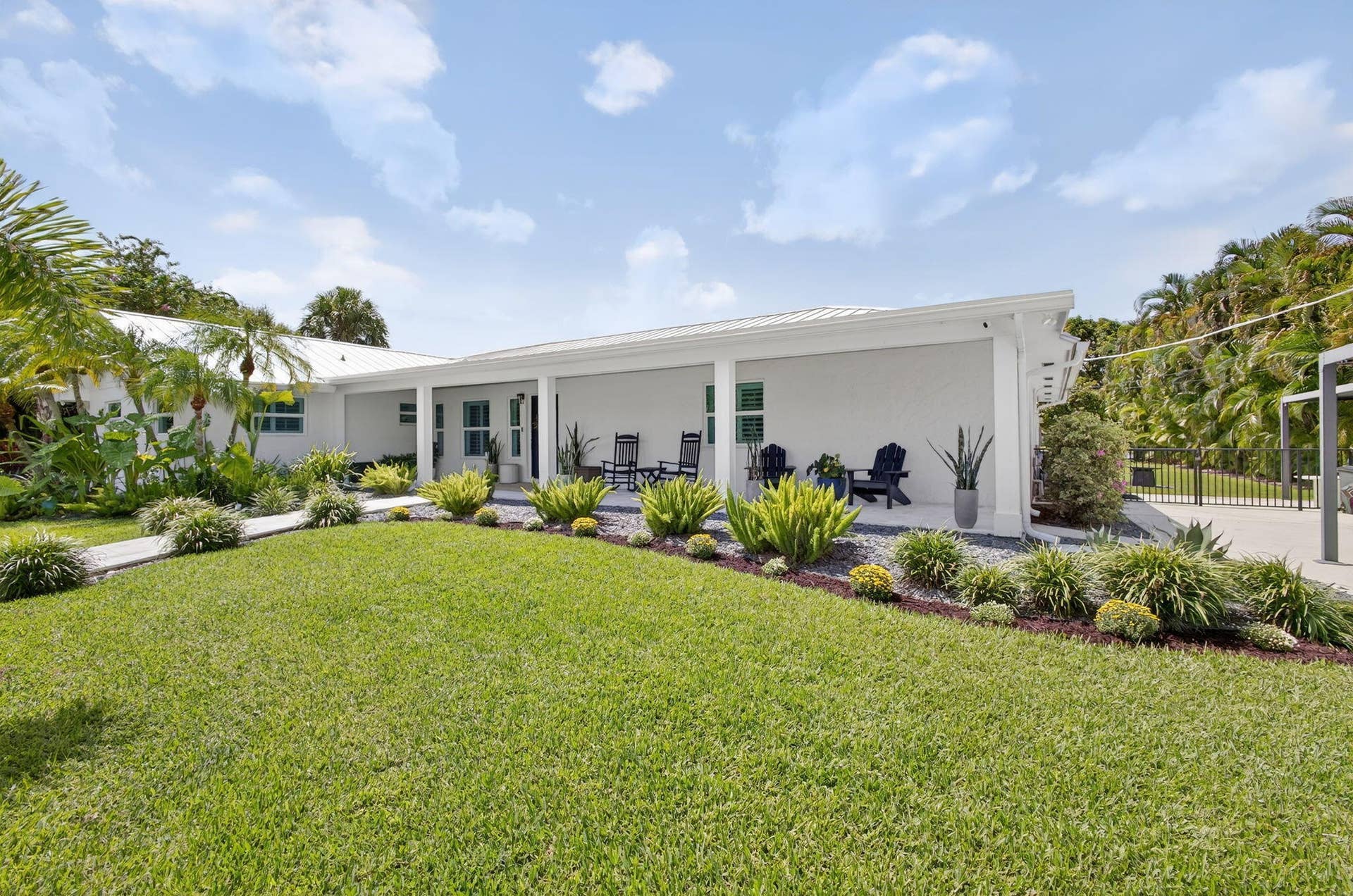 4628 Square Lake Drive, Palm Beach Gardens, FL 33418 Exterior featuring a covered porch, white stucco finish, metal roof, and landscaped lawn with mature greenery