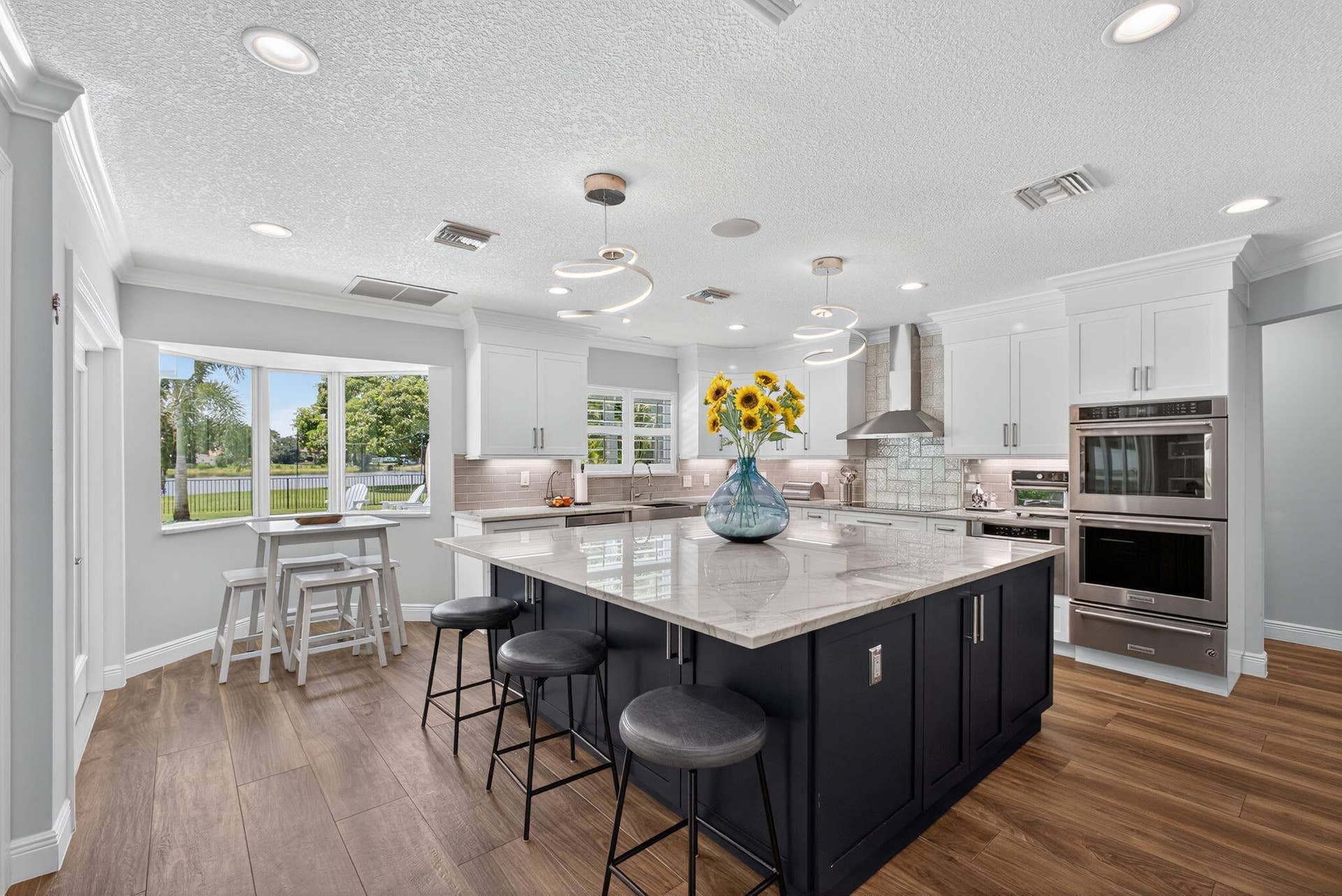 4628 Square Lake Drive, Palm Beach Gardens, FL 33418 Kitchen featuring a contrasting dark base cabinet island with a stone countertop, white upper cabinetry, built-in stainless steel double ovens, recessed ceiling lighting, and wood-finish flooring