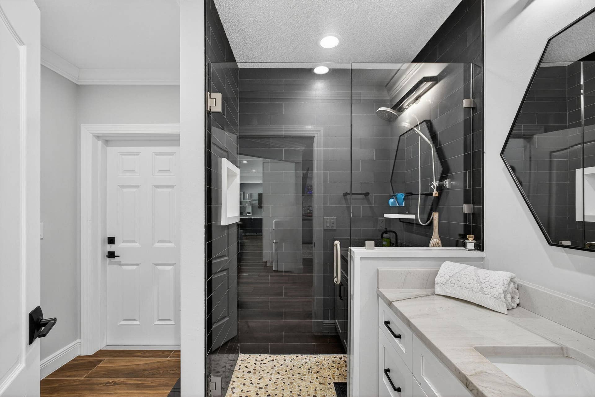 4628 Square Lake Drive, Palm Beach Gardens, FL 33418 Bathroom featuring a walk-in shower with charcoal subway tile, pebble-stone flooring, frameless glass enclosure, white cabinetry with stone-finish countertop, and a geometric-framed wall mirror