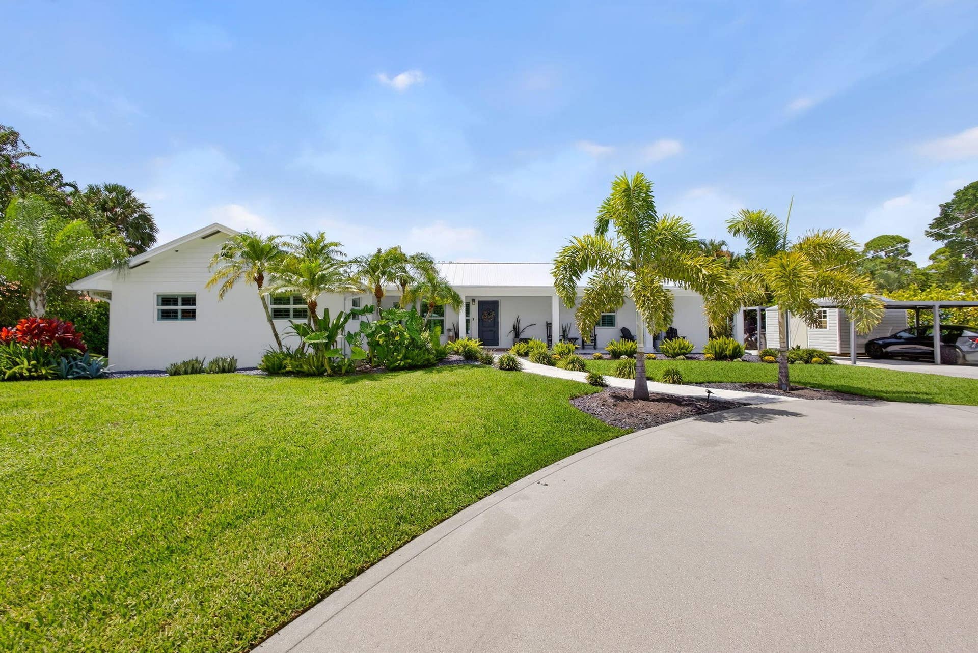 4628 Square Lake Drive, Palm Beach Gardens, FL 33418 Single-story exterior featuring white siding, a metal roof, a paved driveway, a covered carport, and mature palm trees