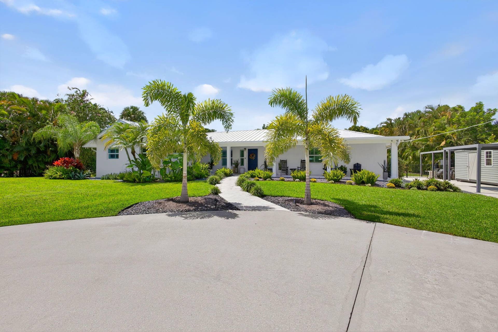 4628 Square Lake Drive, Palm Beach Gardens, FL 33418 Single-story exterior featuring white masonry walls, a metal roof, a paved walkway, a spacious concrete driveway, and established palm trees