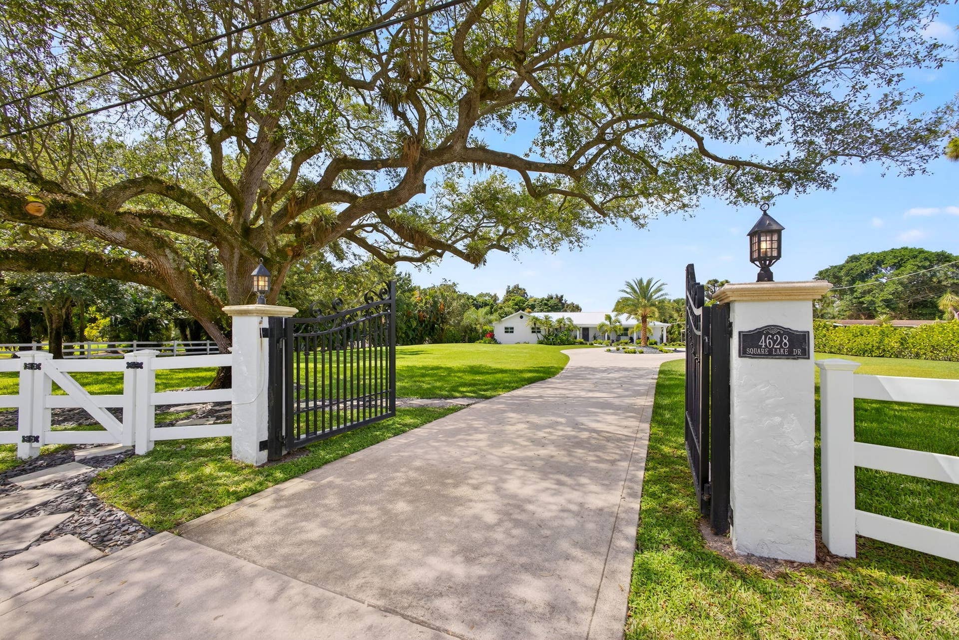 4628 Square Lake Drive, Palm Beach Gardens, FL 33418 Gated entrance featuring decorative metal fencing, concrete-paved driveway, stucco-finished stone pillars, and mounted lantern-style lighting