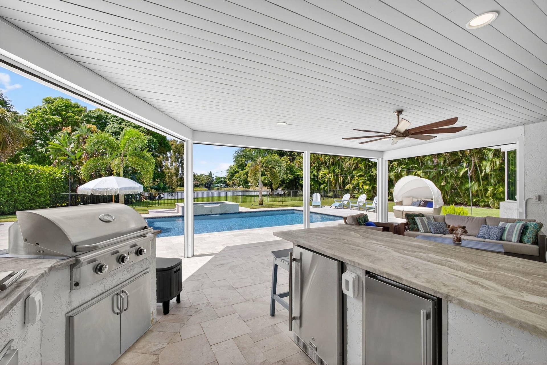 4628 Square Lake Drive, Palm Beach Gardens, FL 33418 Covered patio featuring an outdoor kitchen with stainless steel appliances, stone-finish countertops, tile flooring, and a ceiling fan, opening to a swimming pool with a built-in spa