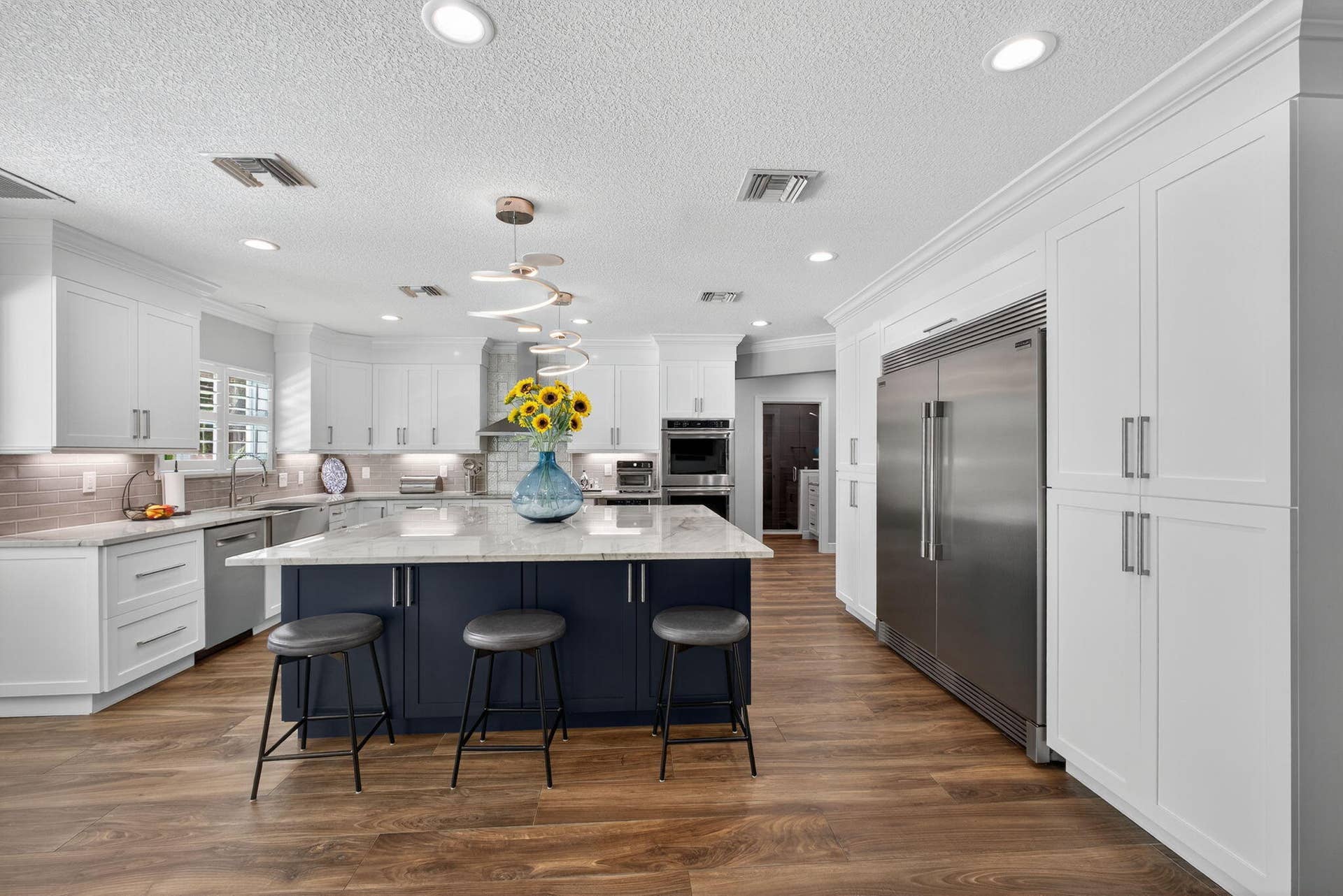 4628 Square Lake Drive, Palm Beach Gardens, FL 33418 Kitchen featuring custom white cabinetry, a contrasting navy blue center island, stone-finish countertops, stainless steel appliances, and wood-finish flooring