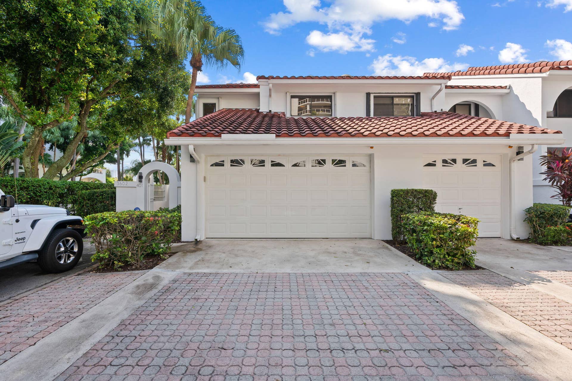 Two-car garage with arched decorative windows, white stucco exterior, terracotta roof tiles, and paver driveway