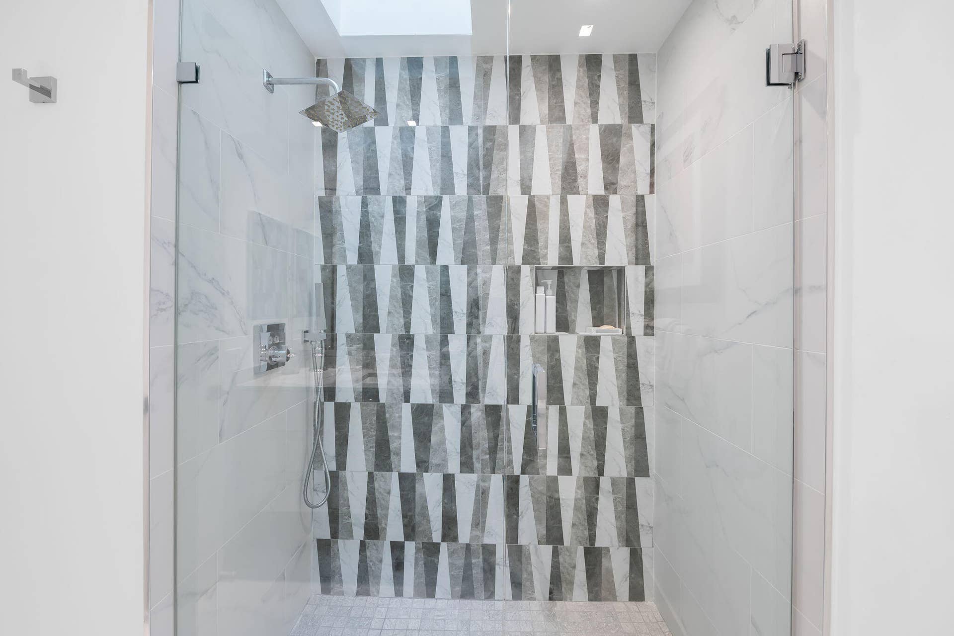 Walk-in shower featuring a frameless glass enclosure, geometric patterned accent tile, built-in recessed shelving, rainfall showerhead, and a skylight