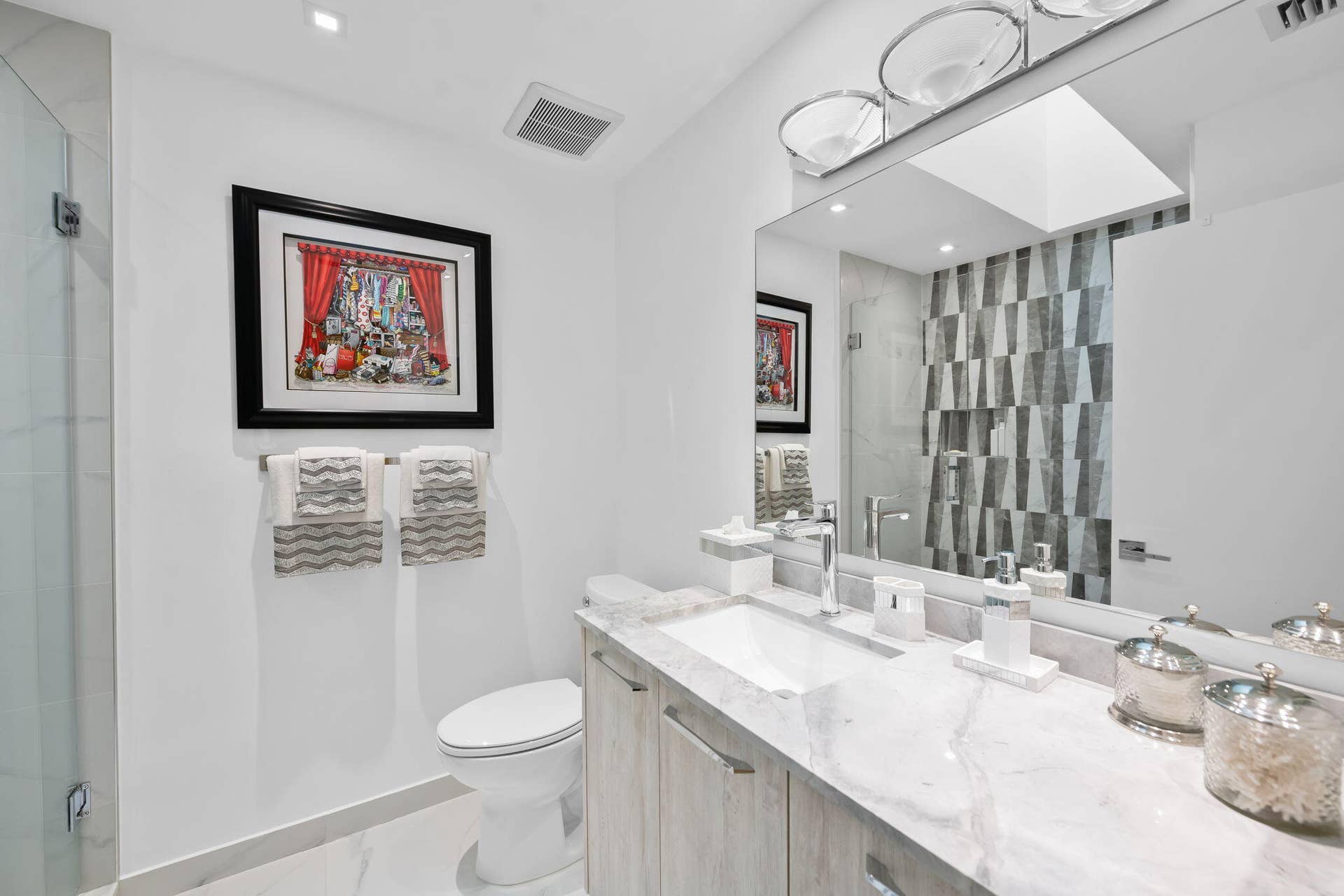 Vanity featuring a light-toned wood-finish cabinet and stone-surface countertop with an integrated basin, large wall-mounted mirror, geometric tile shower surround, and glass-enclosed shower
