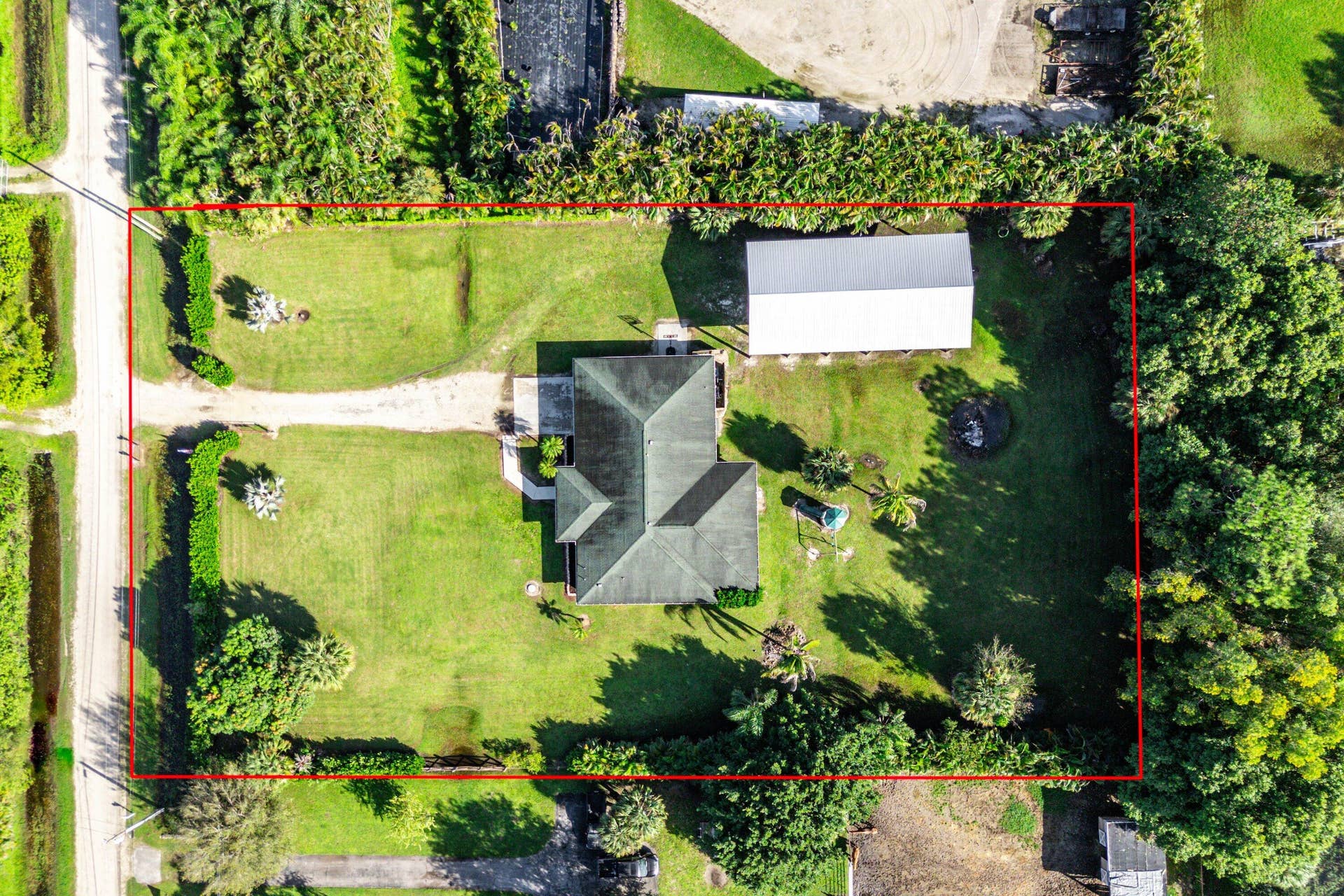 Aerial view of a sprawling lot featuring a detached structure with a metal roof, expansive grass grounds, mature trees, and a gravel driveway