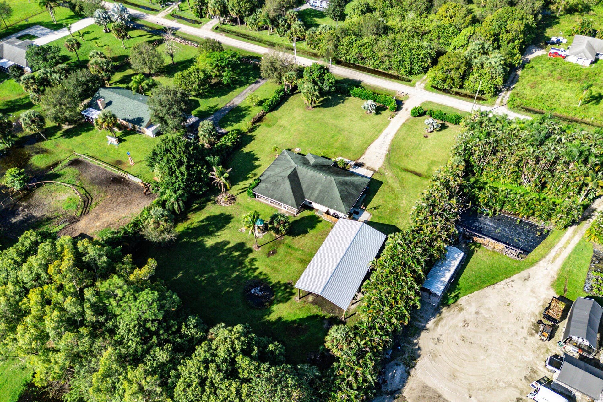 Aerial perspective of a multi-structure property featuring a residence with a dark-tone shingled roof, a detached metal-roofed structure, extensive grassy acreage, mature perimeter trees, and a gravel driveway